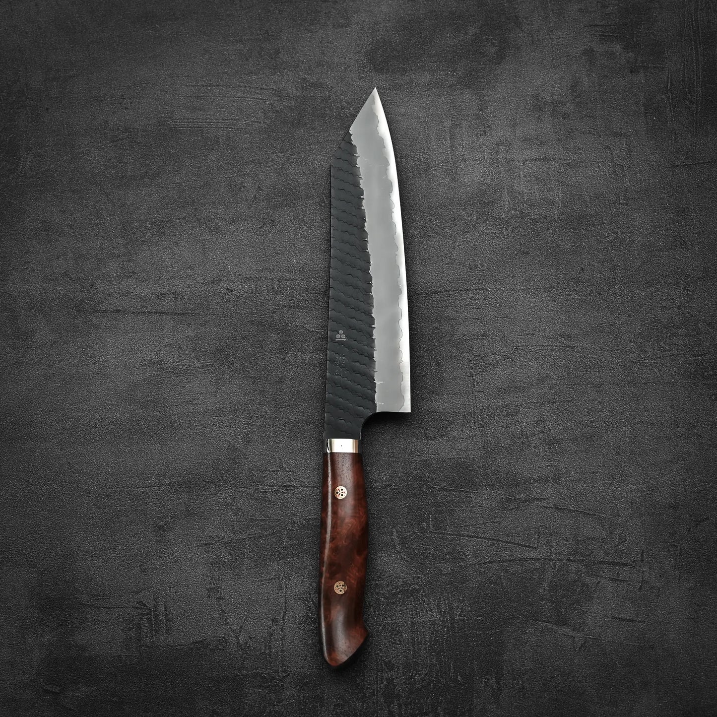 The Nigara Hamono Nigara tsuchime kurouchi SG2 kiritsuke gyuto 210mm, featuring a dark wooden handle and a textured, partially reflective blade, lies flat on a dark, textured surface.