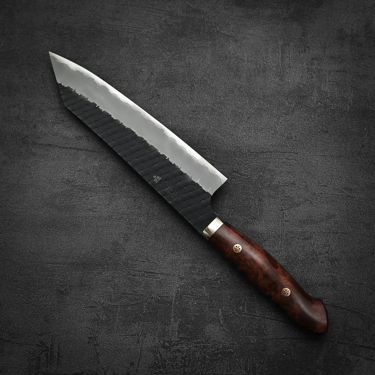 The Nigara Hamono tsuchime kurouchi SG2 kiritsuke gyuto 210mm, featuring a dark, textured blade and polished wooden handle, rests on a dark, textured surface.
