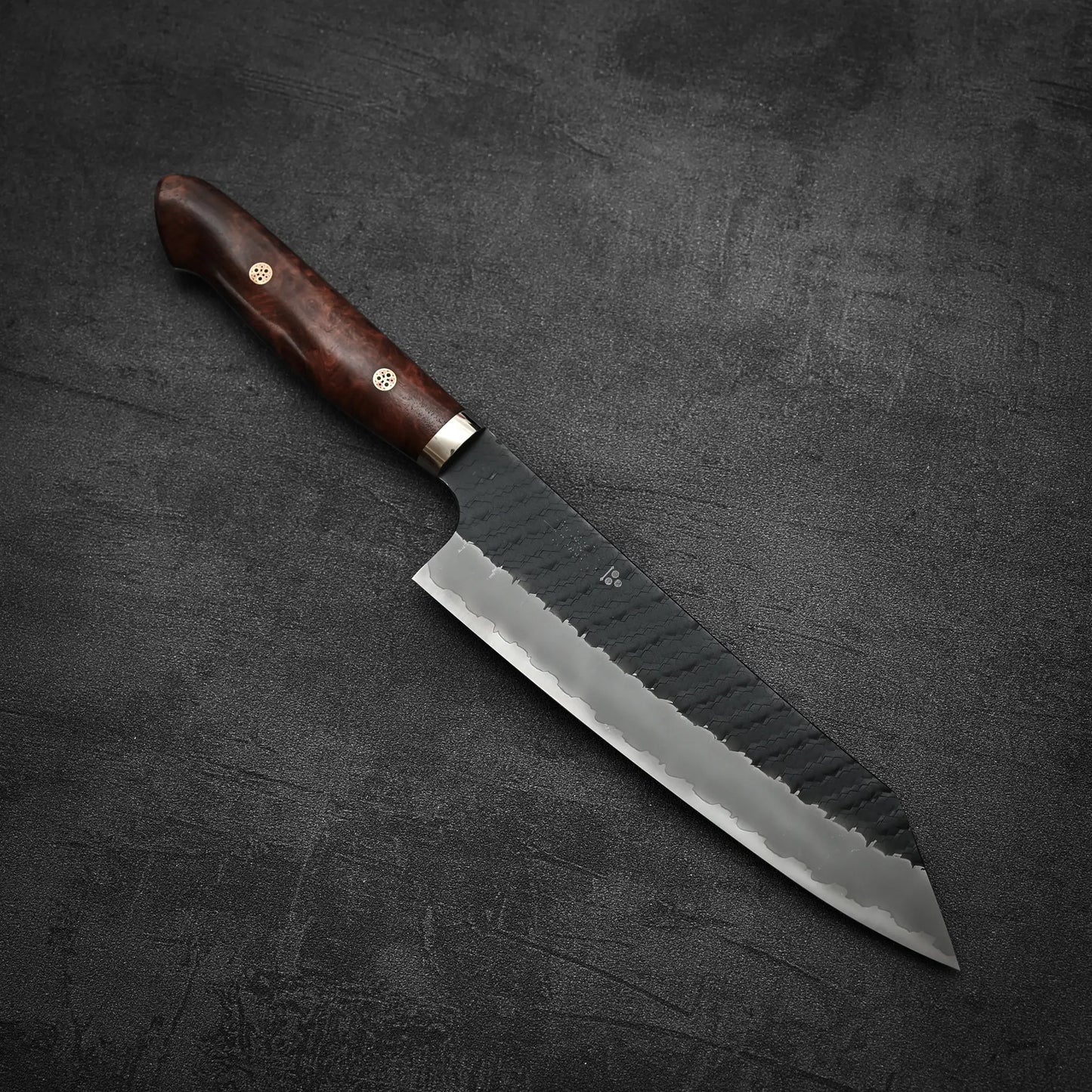 The Nigara Hamono Nigara tsuchime kurouchi SG2 kiritsuke gyuto 210mm, featuring a dark wooden handle and hammered blade, rests on a dark gray textured surface.