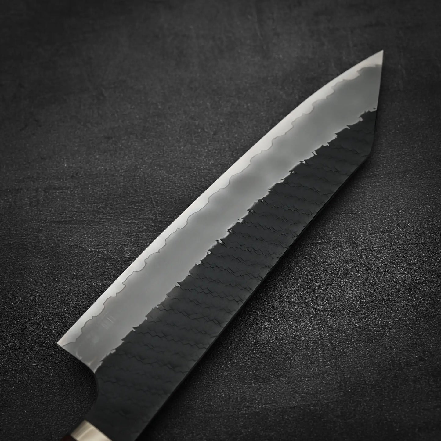 Close-up of the Nigara Hamono Nigara tsuchime kurouchi SG2 kiritsuke gyuto 210mm, featuring a patterned dark handle and a blade with a wavy hamon line, set on a dark textured surface.