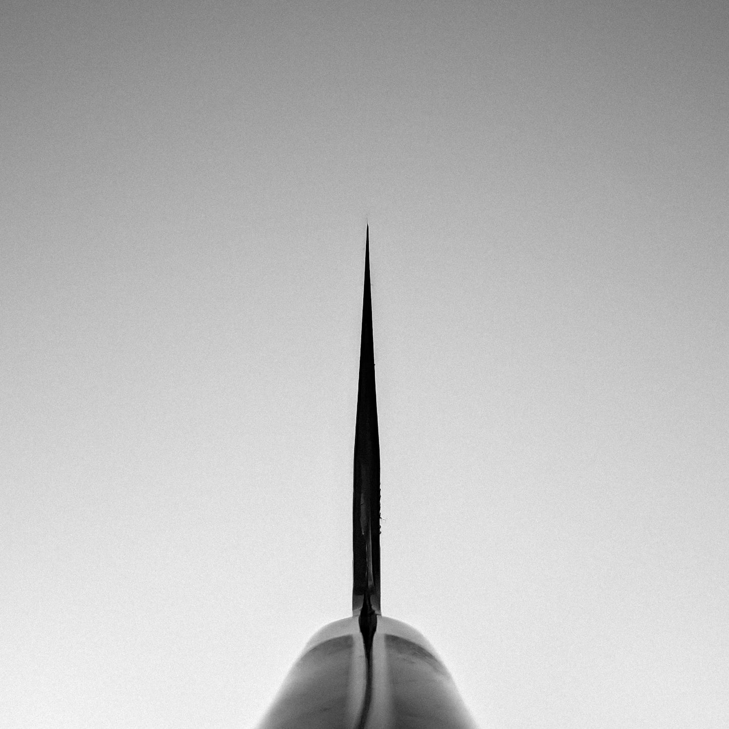 A black and white photo shows the pointed tip of an object, mirroring the sleek symmetry of the Nigara tsuchime kurouchi SG2 kiritsuke gyuto 210mm by Nigara Hamono, vertically centered against a clear sky in a minimalistic, balanced composition.