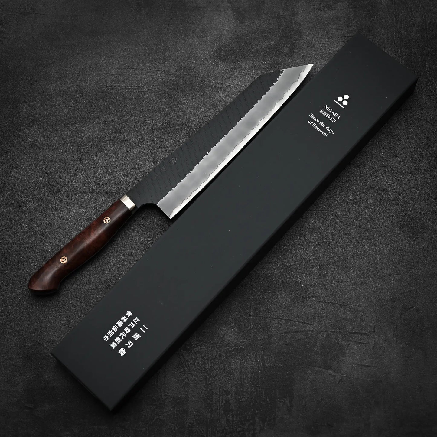 The Nigara Hamono Nigara tsuchime kurouchi SG2 kiritsuke gyuto 240mm, a serrated kitchen knife with a wooden handle, lies beside a black box with Japanese and English text on a dark textured surface.