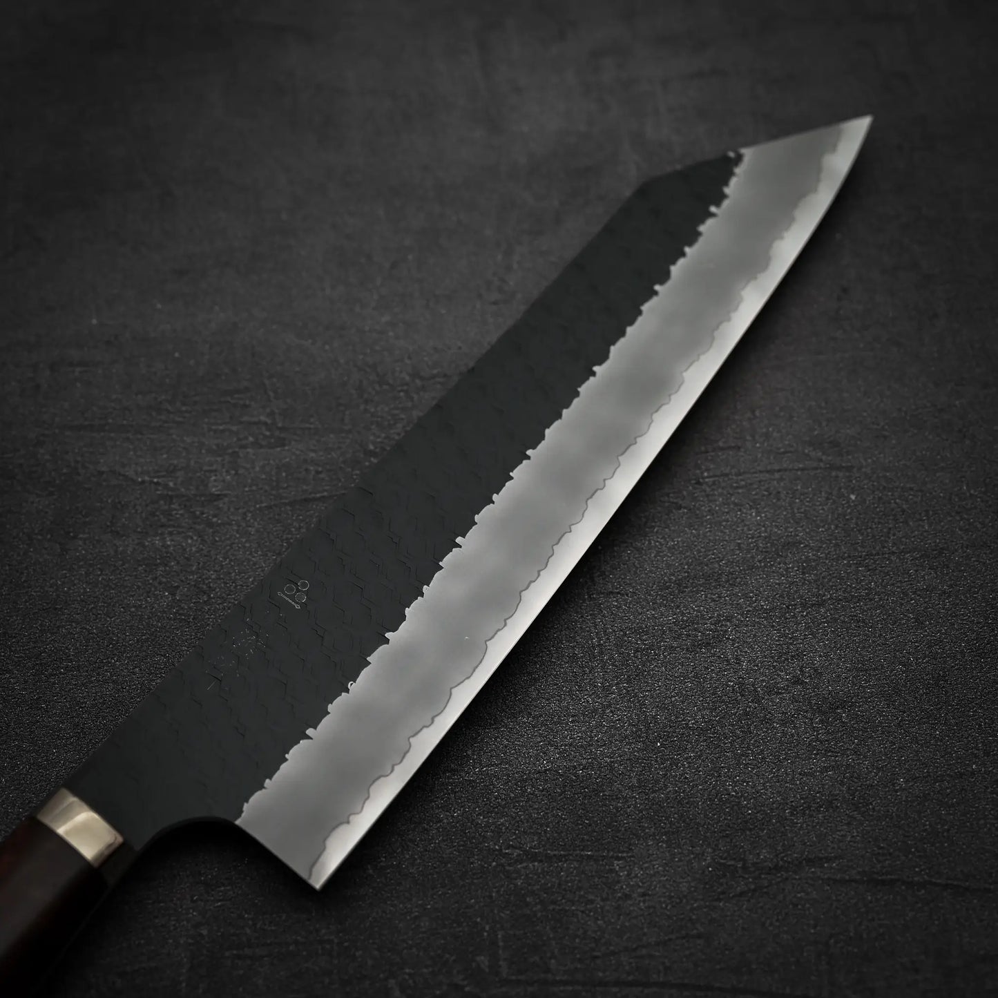 A Nigara Hamono Nigara tsuchime kurouchi SG2 kiritsuke gyuto 240mm with a dark, hammer-patterned stainless steel blade and sharp edge lies on a textured dark surface; its handle is partially visible.