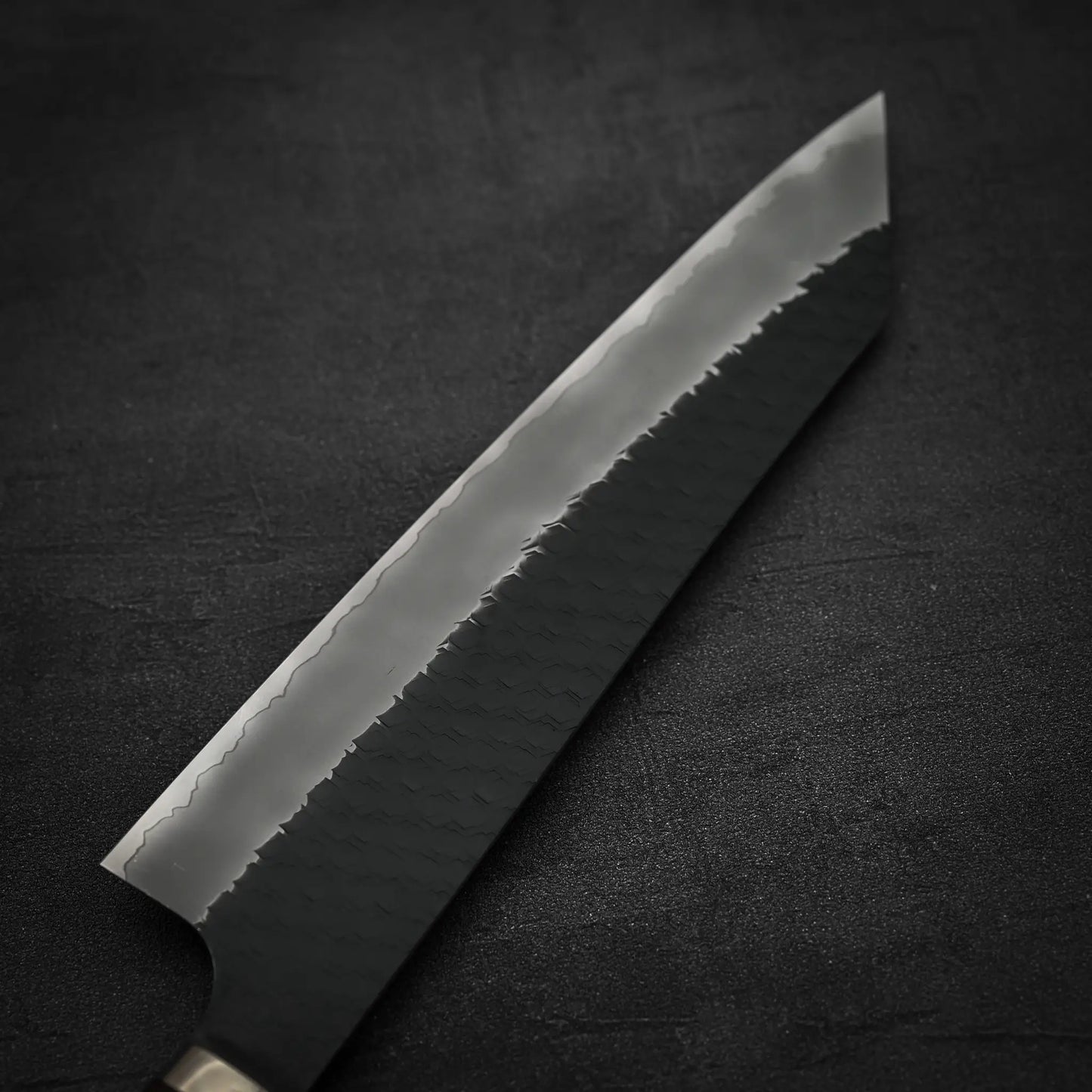A close-up of the Nigara Hamono Nigara tsuchime kurouchi SG2 kiritsuke gyuto 240mm shows its textured blade and razor-sharp edge against a dark background.