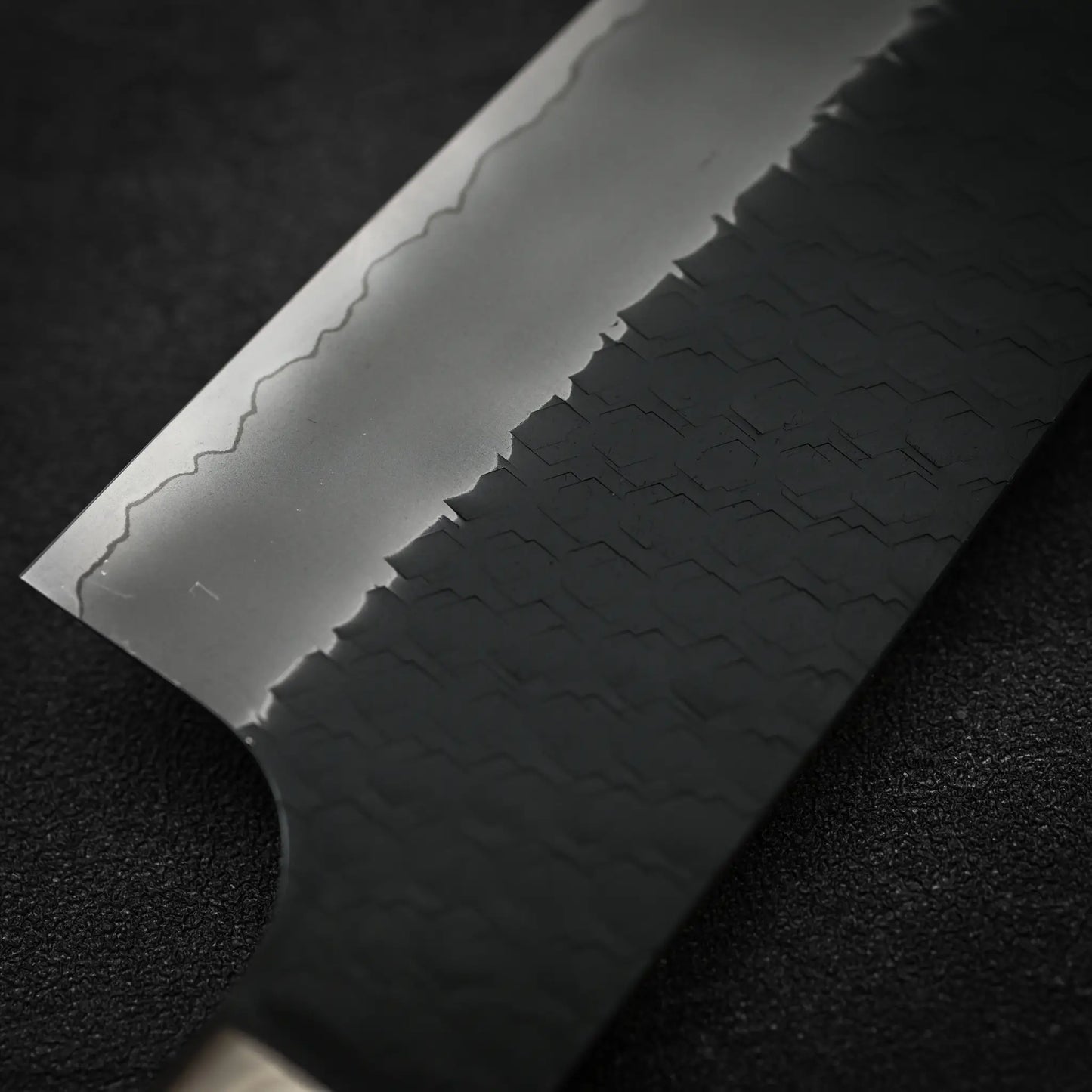Close-up of the Nigara Hamono Nigara tsuchime kurouchi SG2 kiritsuke gyuto 240mm, showing a tsuchime kurouchi finish on the dark lower blade and a polished upper section. The knife rests on a black textured surface.