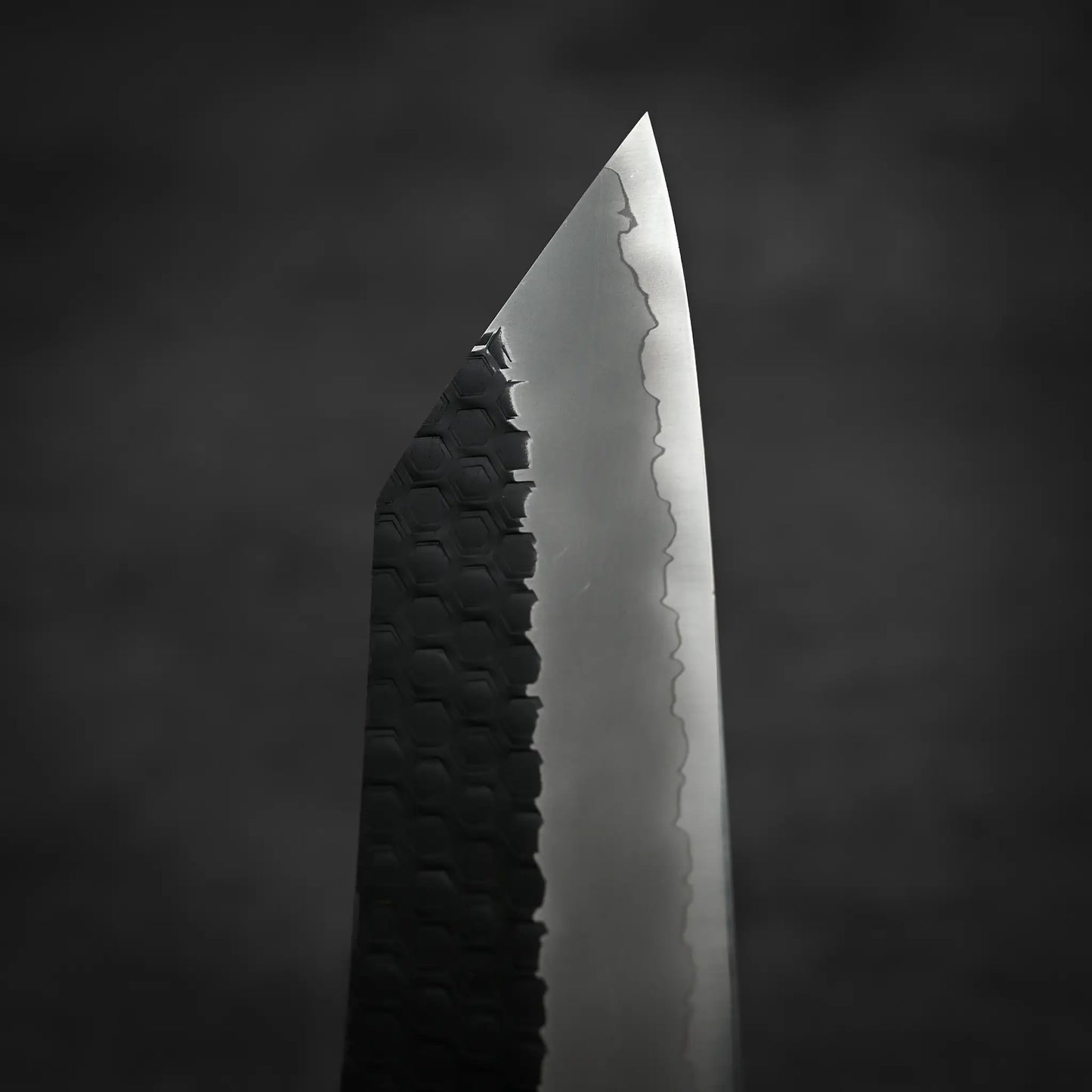A close-up of the Nigara Hamono Nigara tsuchime kurouchi SG2 kiritsuke gyuto 240mm shows its patterned SG2 stainless steel blade and sharp pointed tip against a dark, blurred background.