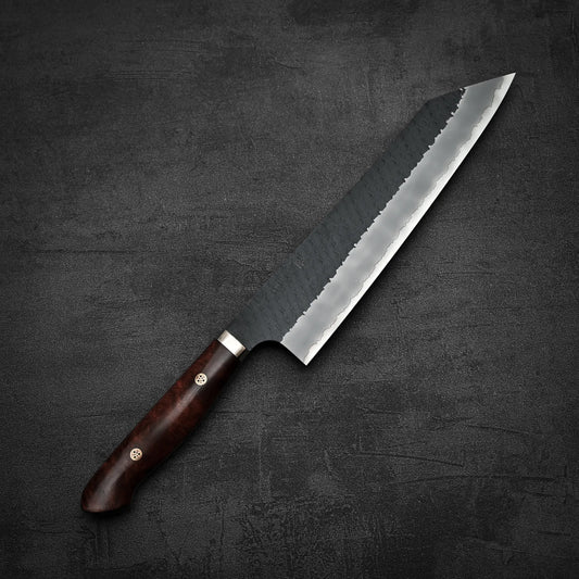 The Nigara Hamono Nigara tsuchime kurouchi SG2 kiritsuke gyuto 240mm, a Japanese-style chef’s knife with a dark wooden handle and hammered SG2 blade, is displayed on a dark textured surface.