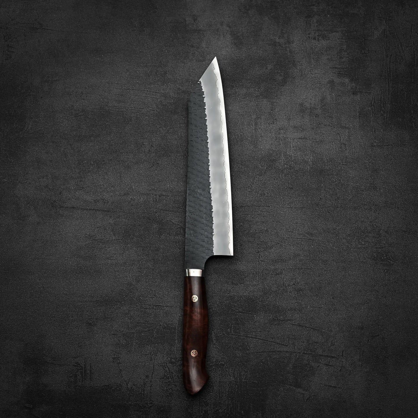 The Nigara Hamono Nigara tsuchime kurouchi SG2 kiritsuke gyuto 240mm, with its dark wooden handle and hammered blade, rests elegantly on a dark, textured surface.