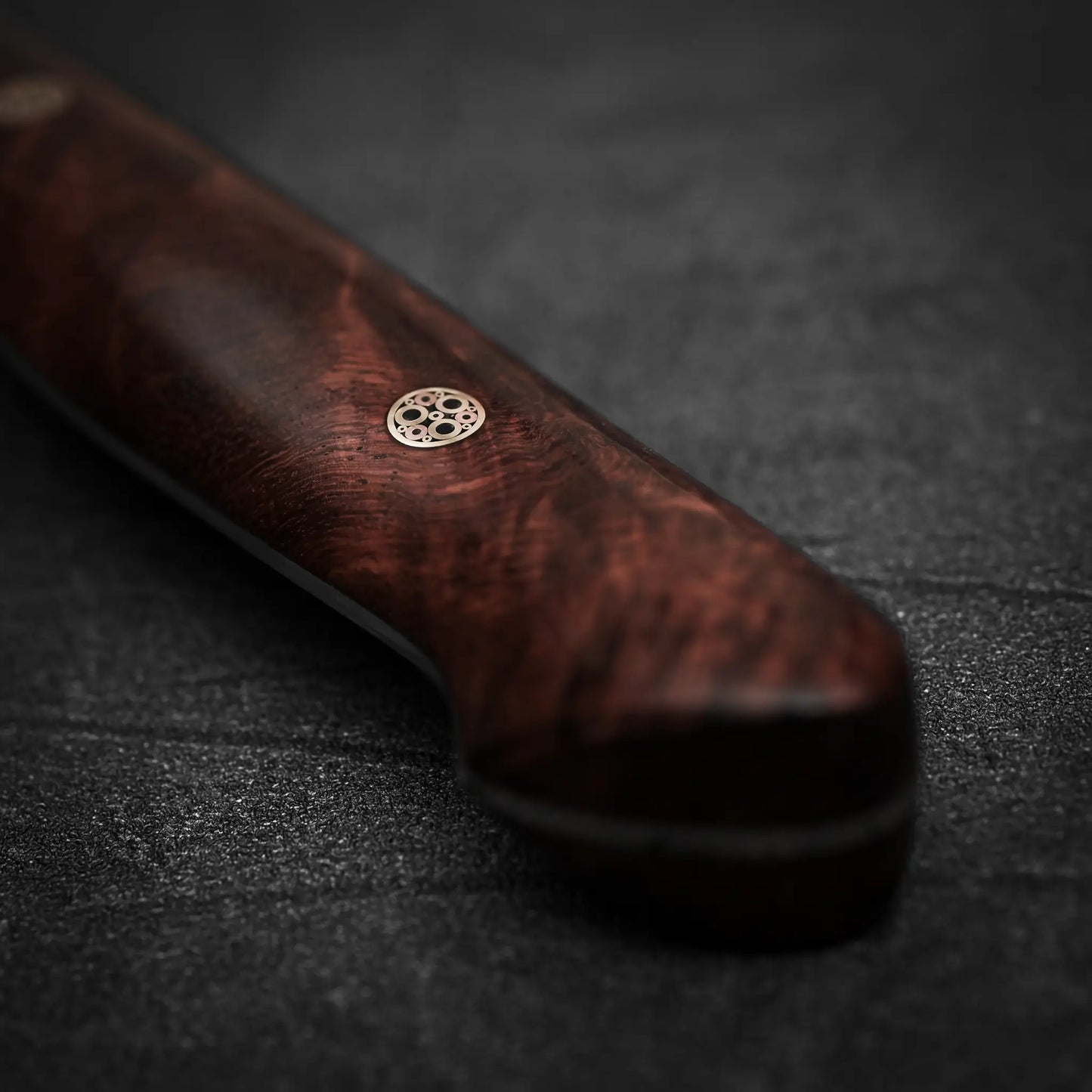 Close-up of the dark wooden handle with a decorative metal pin of the Nigara Hamono Nigara tsuchime kurouchi SG2 kiritsuke gyuto 240mm, resting on a textured black surface; artisan details are visible, though the blade is out of frame.
