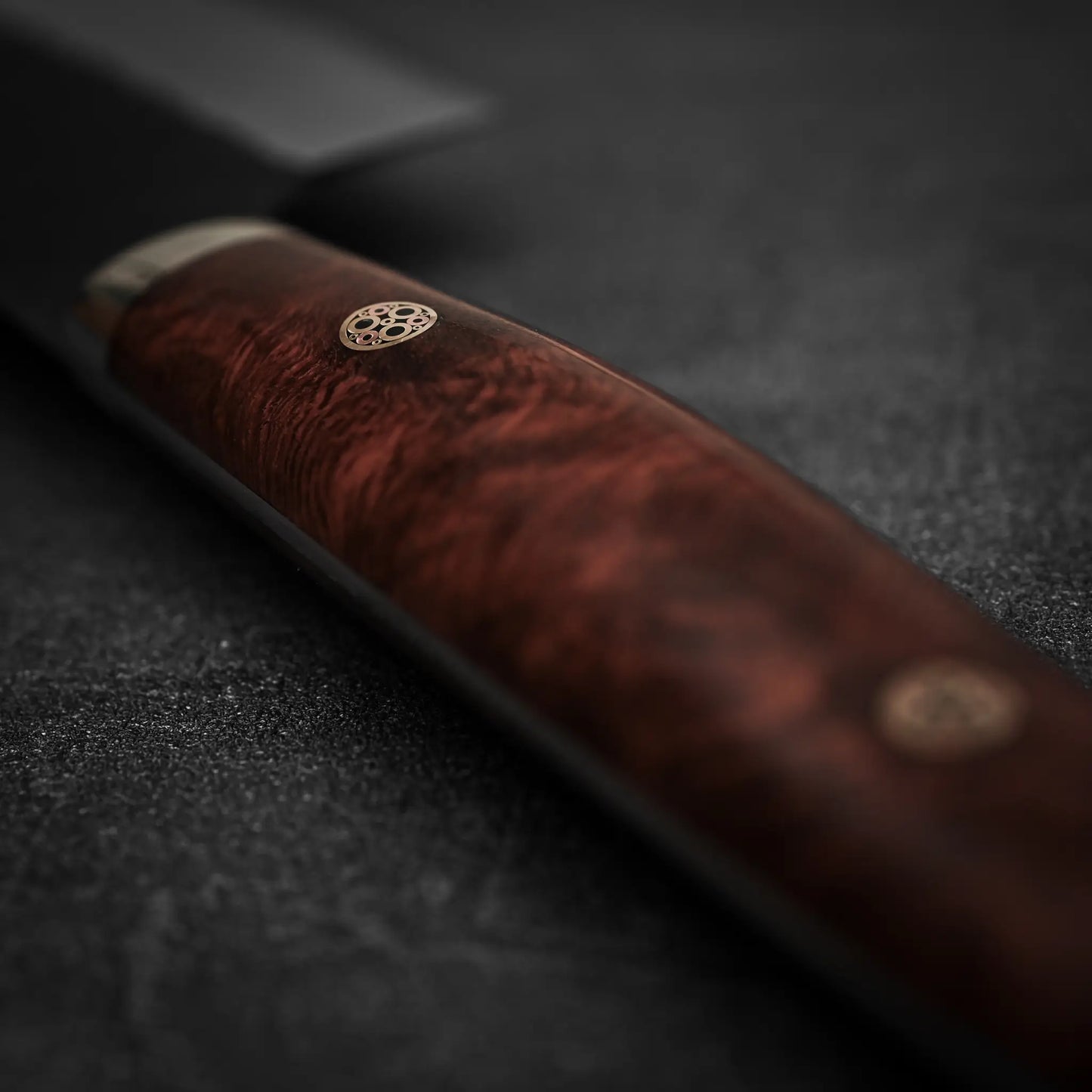 Close-up of the Nigara Hamono Nigara tsuchime kurouchi SG2 kiritsuke gyuto 240mm handle, crafted from polished reddish-brown wood with metal accents, resting on a dark surface. The blade showcases premium SG2 stainless steel craftsmanship.