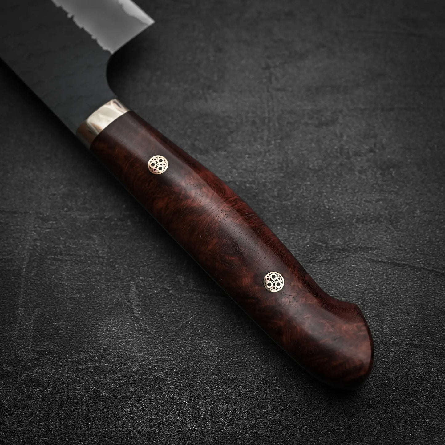 Close-up of the Nigara Hamono Nigara tsuchime kurouchi SG2 kiritsuke gyuto 240mm, showing its polished dark wood handle with two metal rivets and a partially visible SG2 stainless steel blade against a textured dark background.