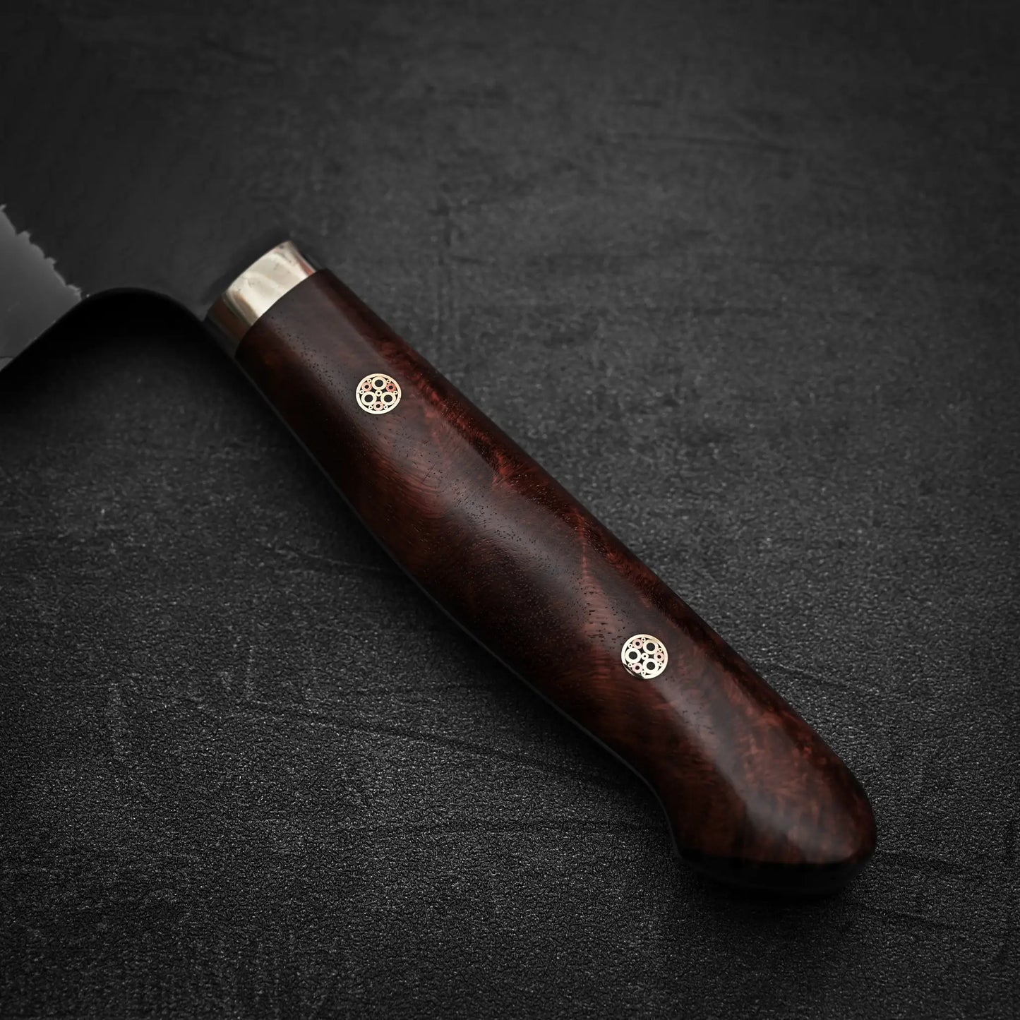 Close-up of the Nigara Hamono Nigara tsuchime kurouchi SG2 kiritsuke gyuto 240mm: a dark wooden handle with two decorative rivets rests on a textured black surface; the SG2 stainless steel blade is partially visible on the left.