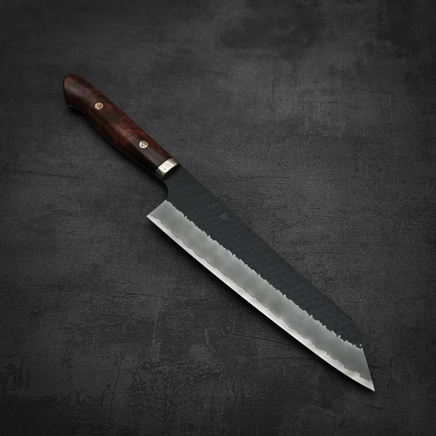 The Nigara Hamono Nigara tsuchime kurouchi SG2 kiritsuke gyuto 240mm, featuring a hammered finish and dark wooden handle, rests on a textured dark gray surface.