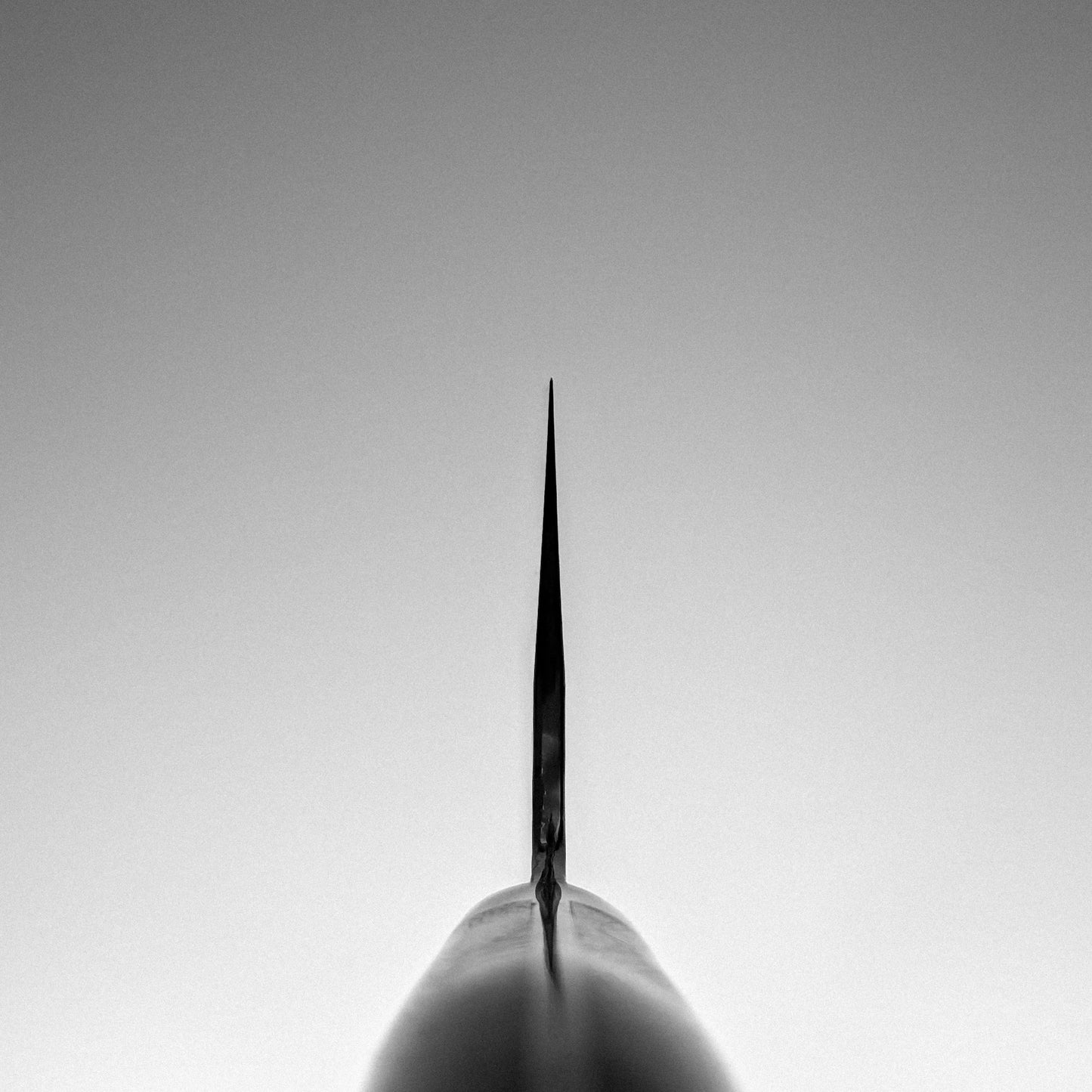 A black and white photo captures the base of a pointed, vertical structure resembling the Nigara Hamono Nigara tsuchime kurouchi SG2 kiritsuke gyuto 240mm blade, centered against a clear, cloudless sky.