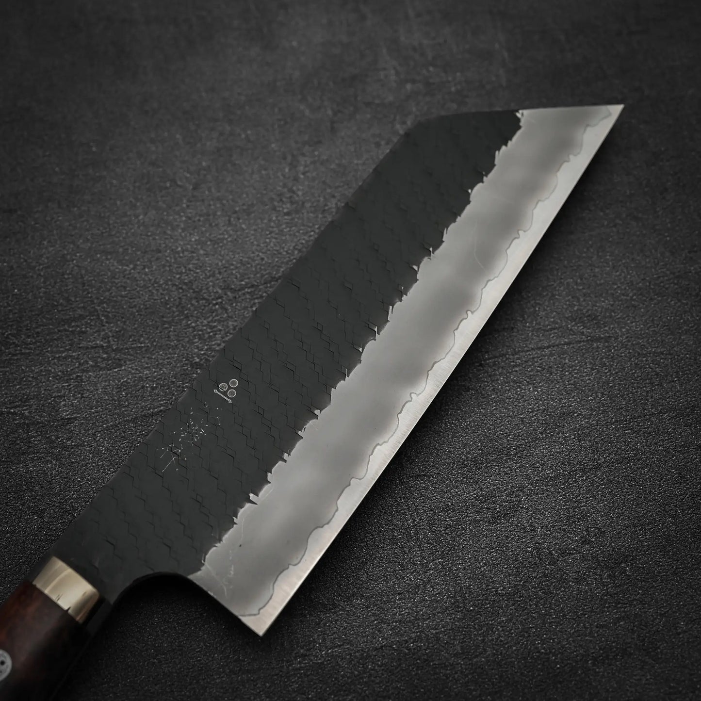 A close-up of the Nigara Hamono Nigara tsuchime kurouchi SG2 kiritsuke nakiri 180mm, featuring a dark handle and patterned steel blade with a wavy edge and tsuchime kurouchi finish, placed on a textured dark gray surface.