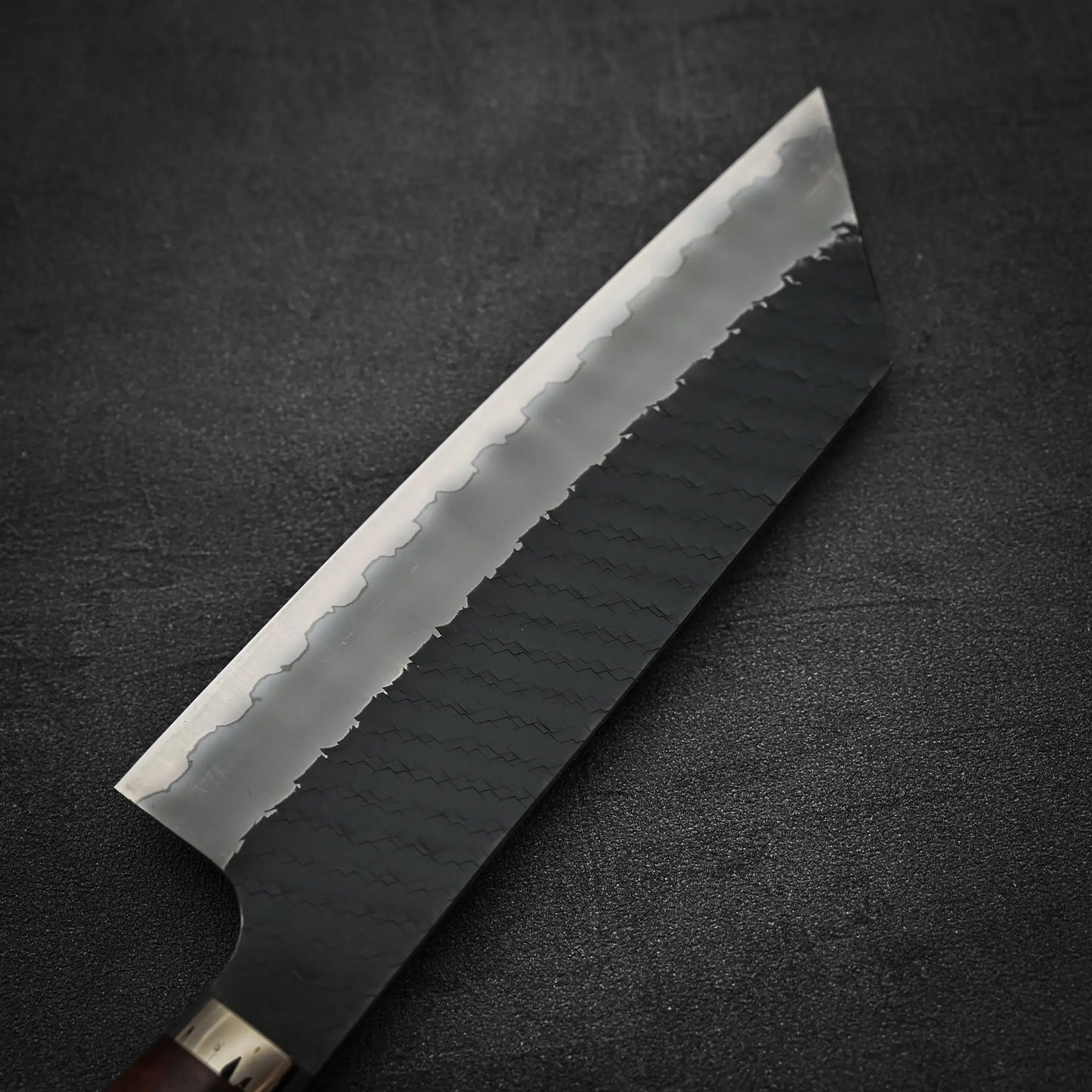 A close-up of the Nigara Hamono Nigara tsuchime kurouchi SG2 kiritsuke nakiri 180mm, featuring a tsuchime kurouchi patterned blade and dark handle, resting on a textured black surface.