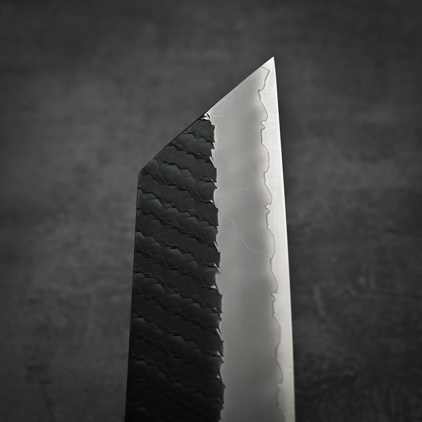 Close-up of the Nigara Hamono Nigara tsuchime kurouchi SG2 kiritsuke nakiri 180mm, highlighting its dark, textured handle and sharply angled, polished blade against a blurred dark background.