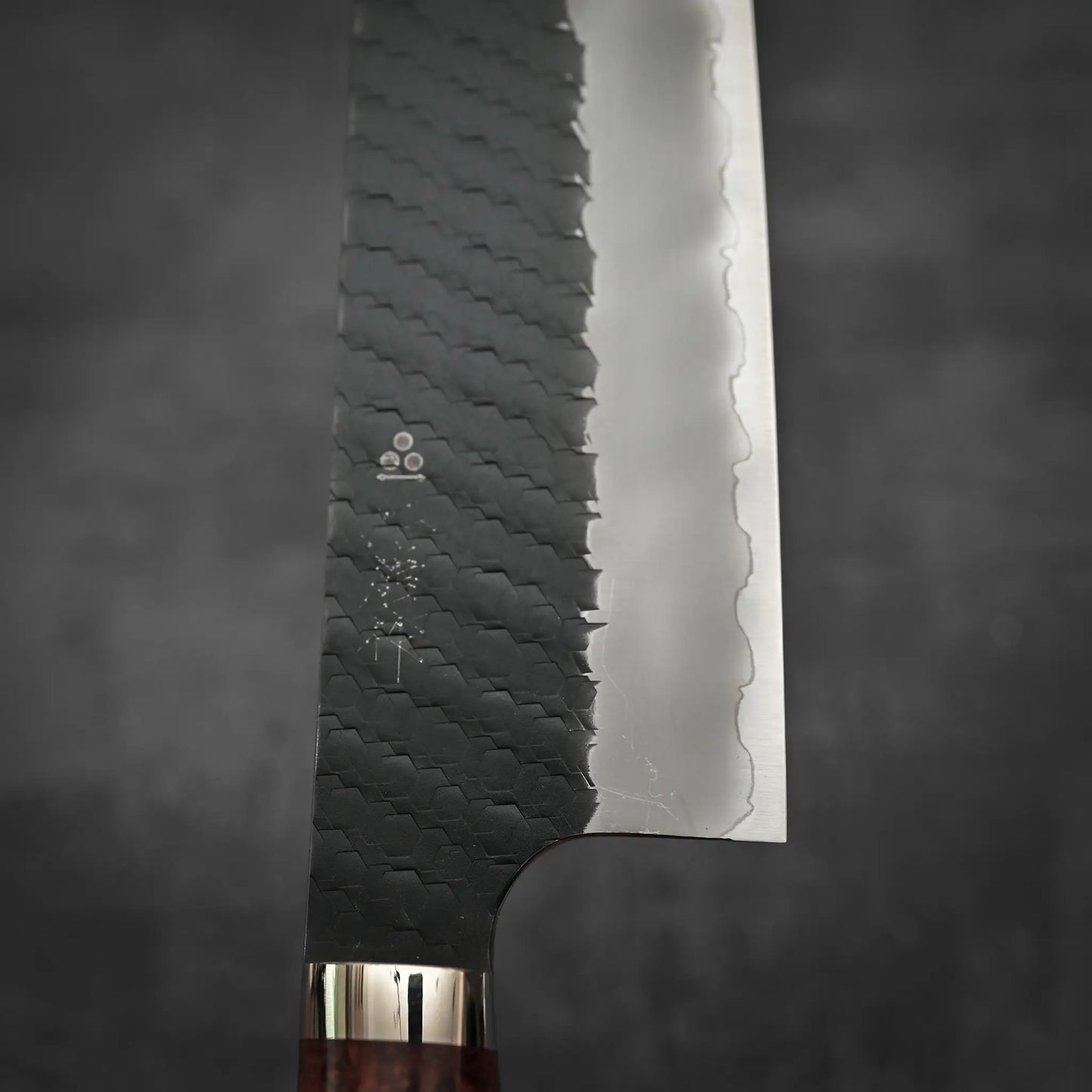 Close-up of the Nigara Hamono Nigara tsuchime kurouchi SG2 kiritsuke nakiri 180mm, showcasing its hammered black finish and polished wavy blade edge, set against a dark, blurred background.