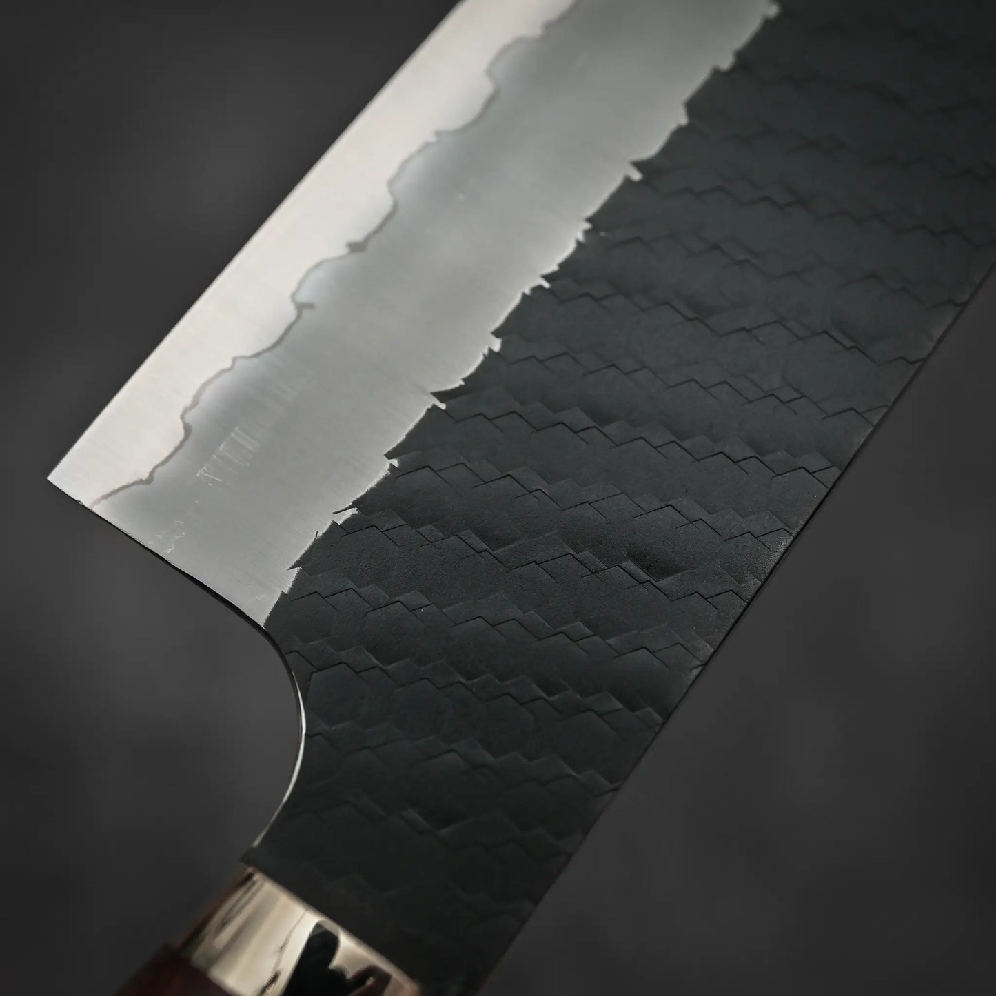 Close-up of the Nigara Hamono Nigara tsuchime kurouchi SG2 kiritsuke nakiri 180mm, showcasing a wavy hamon line and dark Damascus tsuchime kurouchi finish, set against a blurred dark background.