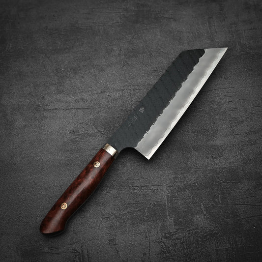 A Nigara Hamono Nigara tsuchime kurouchi SG2 kiritsuke nakiri 180mm with a dark wooden handle and wide gray steel blade rests on a textured dark gray surface.