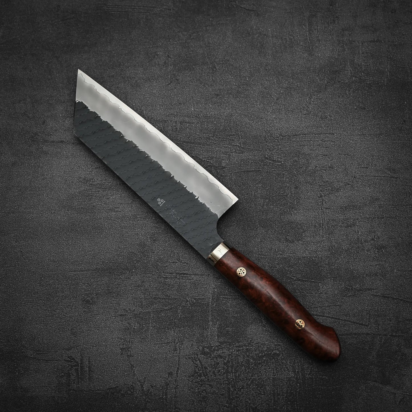 A Nigara Hamono Nigara tsuchime kurouchi SG2 kiritsuke nakiri 180mm with a dark wood handle and brass rivets rests on a textured dark gray surface.