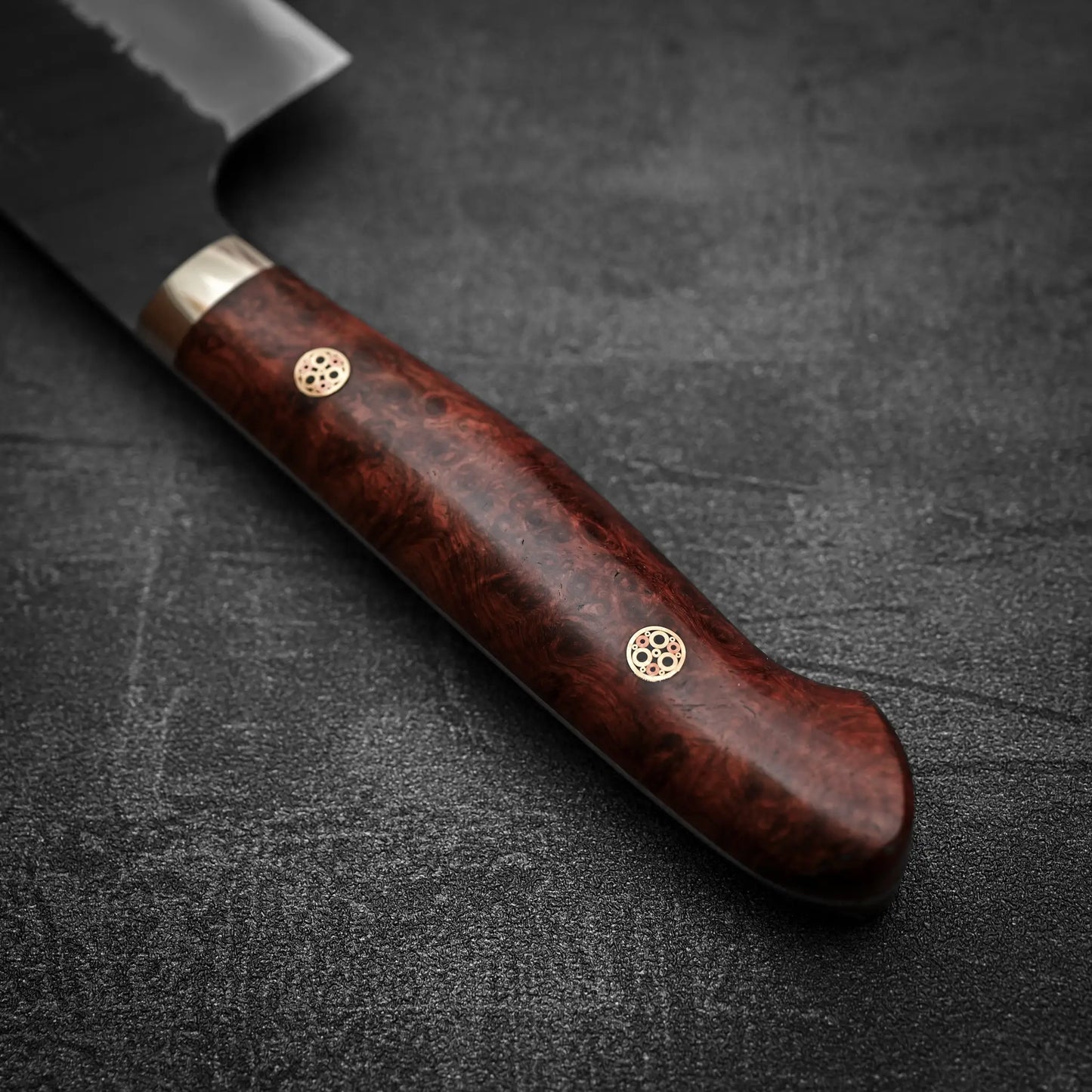 Close-up of the Nigara Hamono Nigara tsuchime kurouchi SG2 kiritsuke nakiri 180mm knife with a polished wooden handle and two decorative rivets, resting on a dark textured surface.