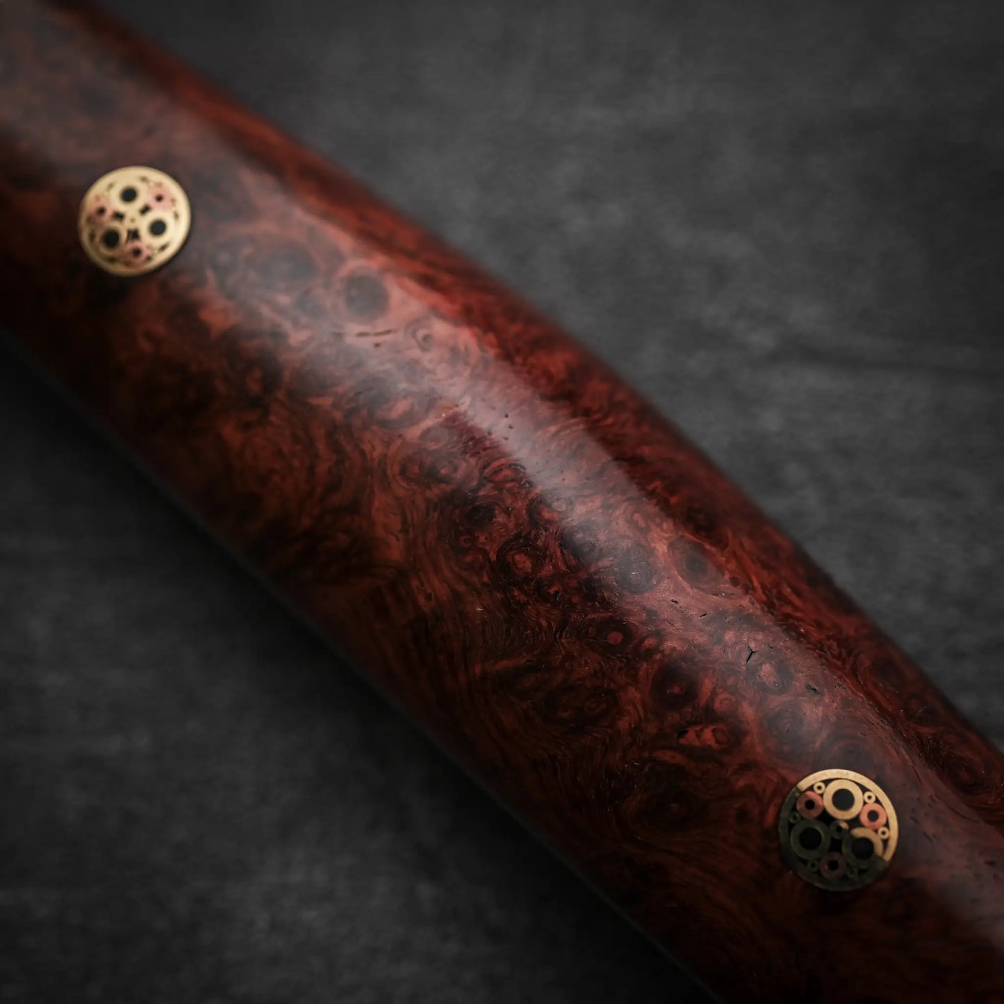 A close-up of the Nigara Hamono Nigara tsuchime kurouchi SG2 kiritsuke nakiri 180mm shows its polished wooden handle with a reddish-brown burl pattern and gear-like metallic inlays, echoing the knifes unique textured finish, set against a dark background.