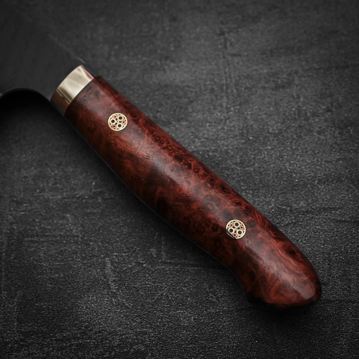 Close-up of the Nigara Hamono Nigara tsuchime kurouchi SG2 kiritsuke nakiri 180mm knife handle, featuring polished dark marbled wood, two decorative pins, and a silver bolster, resting on a dark textured surface.