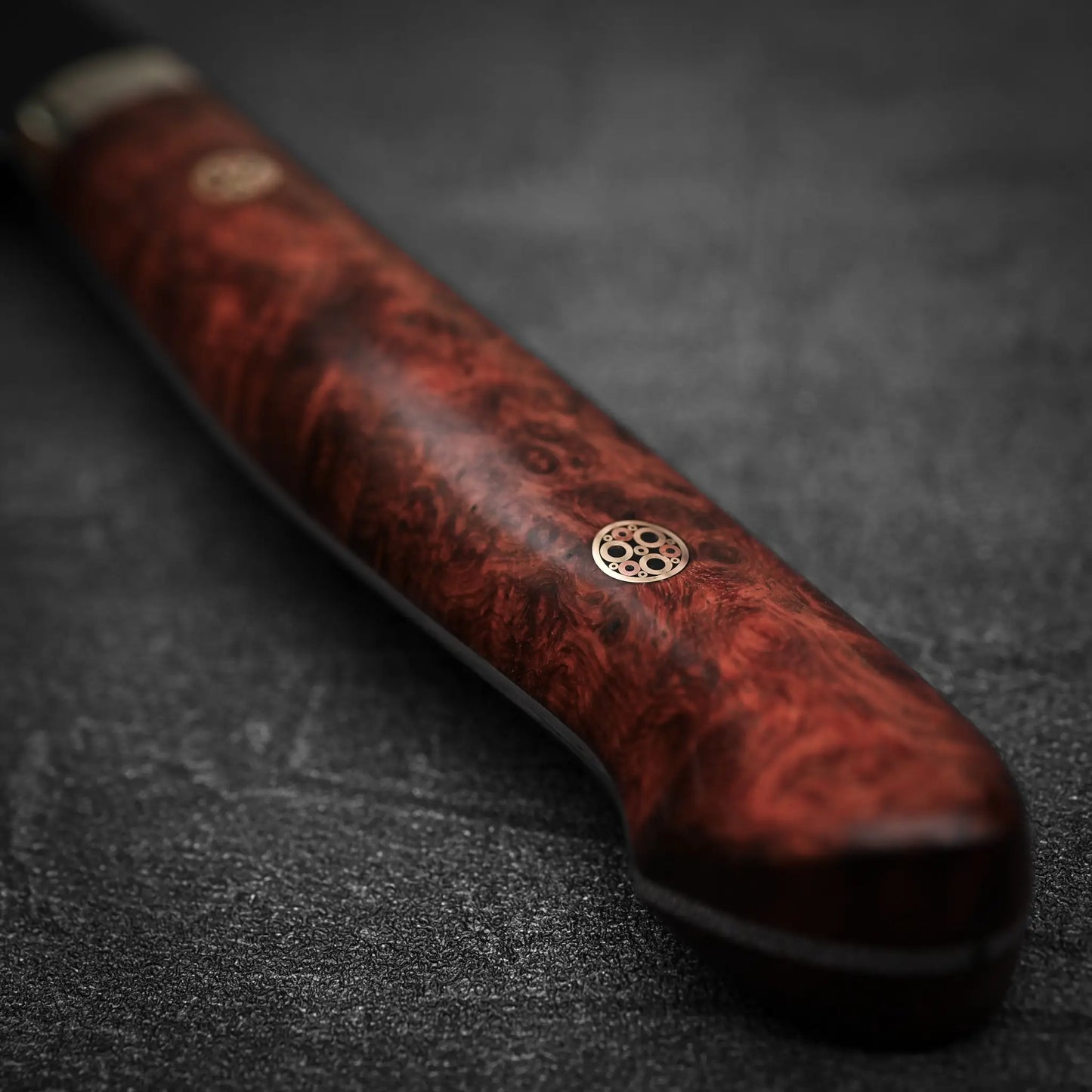 Close-up of the Nigara Hamono Nigara tsuchime kurouchi SG2 kiritsuke nakiri 180mm knife handle, crafted from polished, richly-grained reddish wood with circular metal accents, set against a dark textured background.