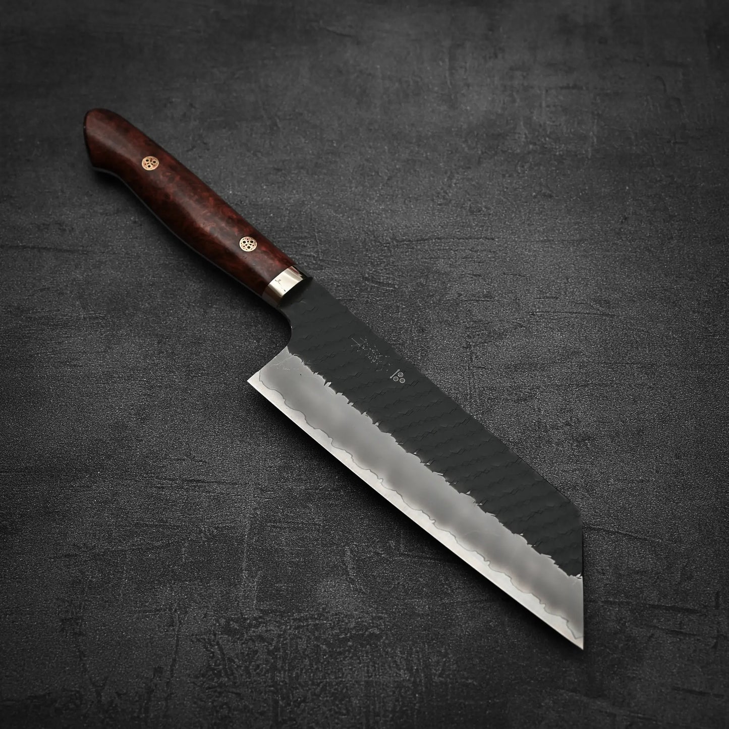 The Nigara Hamono tsuchime kurouchi SG2 kiritsuke nakiri 180mm, with a dark wooden handle and a wide, straight-edged blade, lies on a dark textured surface.