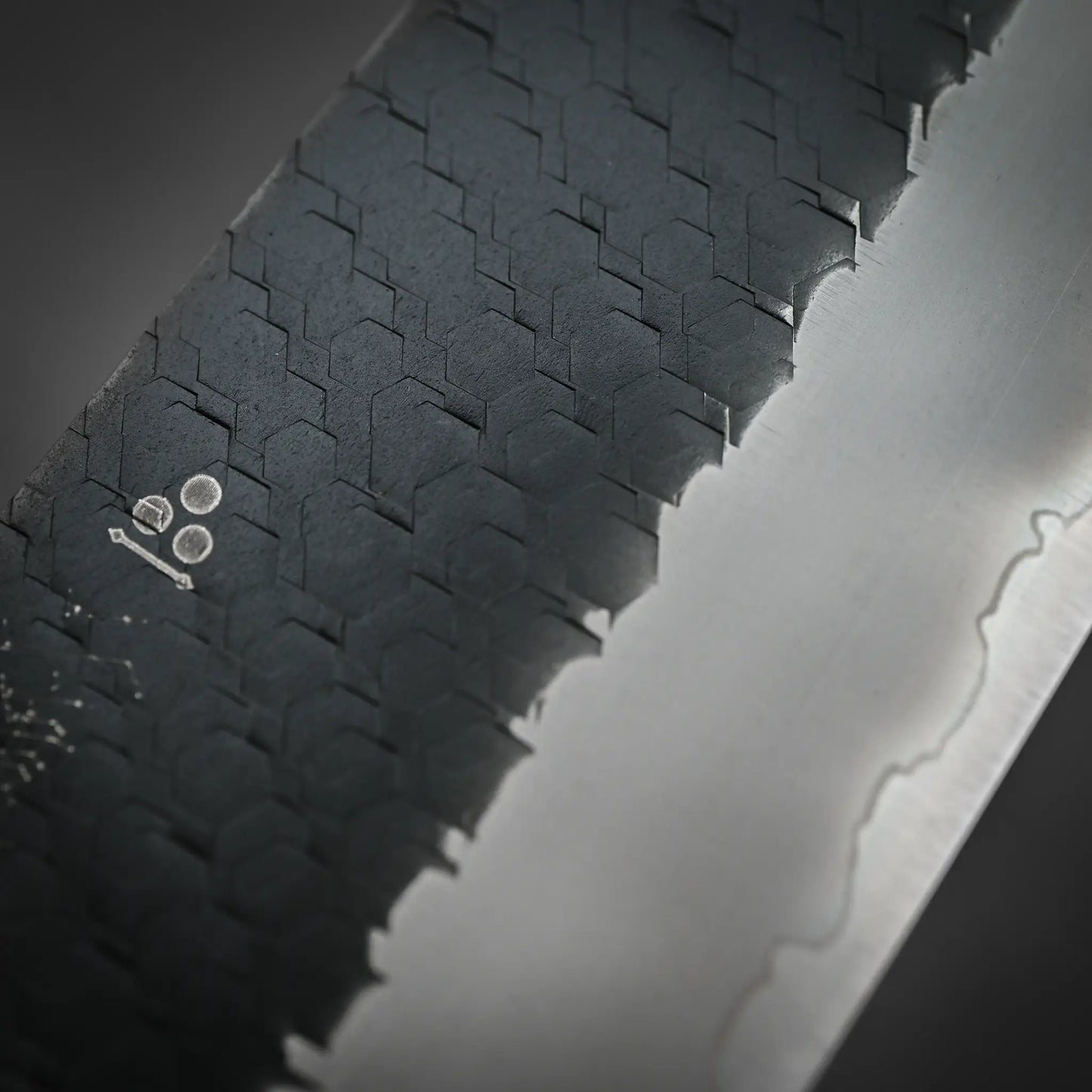 Close-up of the Nigara Hamono Nigara tsuchime kurouchi SG2 kiritsuke sujihiki 255mm blade, showing its SG2 steel edge, wavy line dividing textured dark and smooth light sections, and the maker’s mark engraved on the blade.