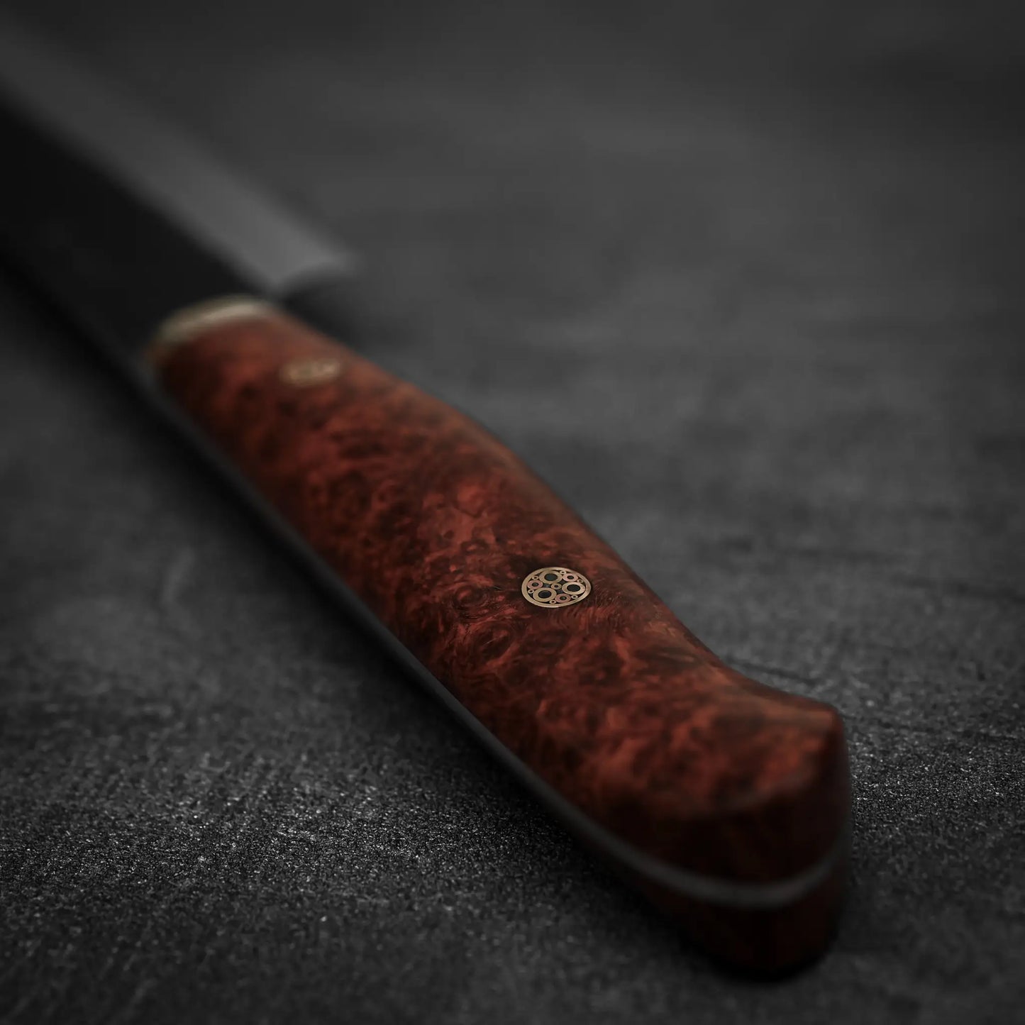 Close-up of a Nigara Hamono Nigara tsuchime kurouchi SG2 kiritsuke sujihiki 255mm, featuring a polished reddish-brown handle with a metal pin. The SG2 stainless steel blade appears partially visible against a dark, textured background.