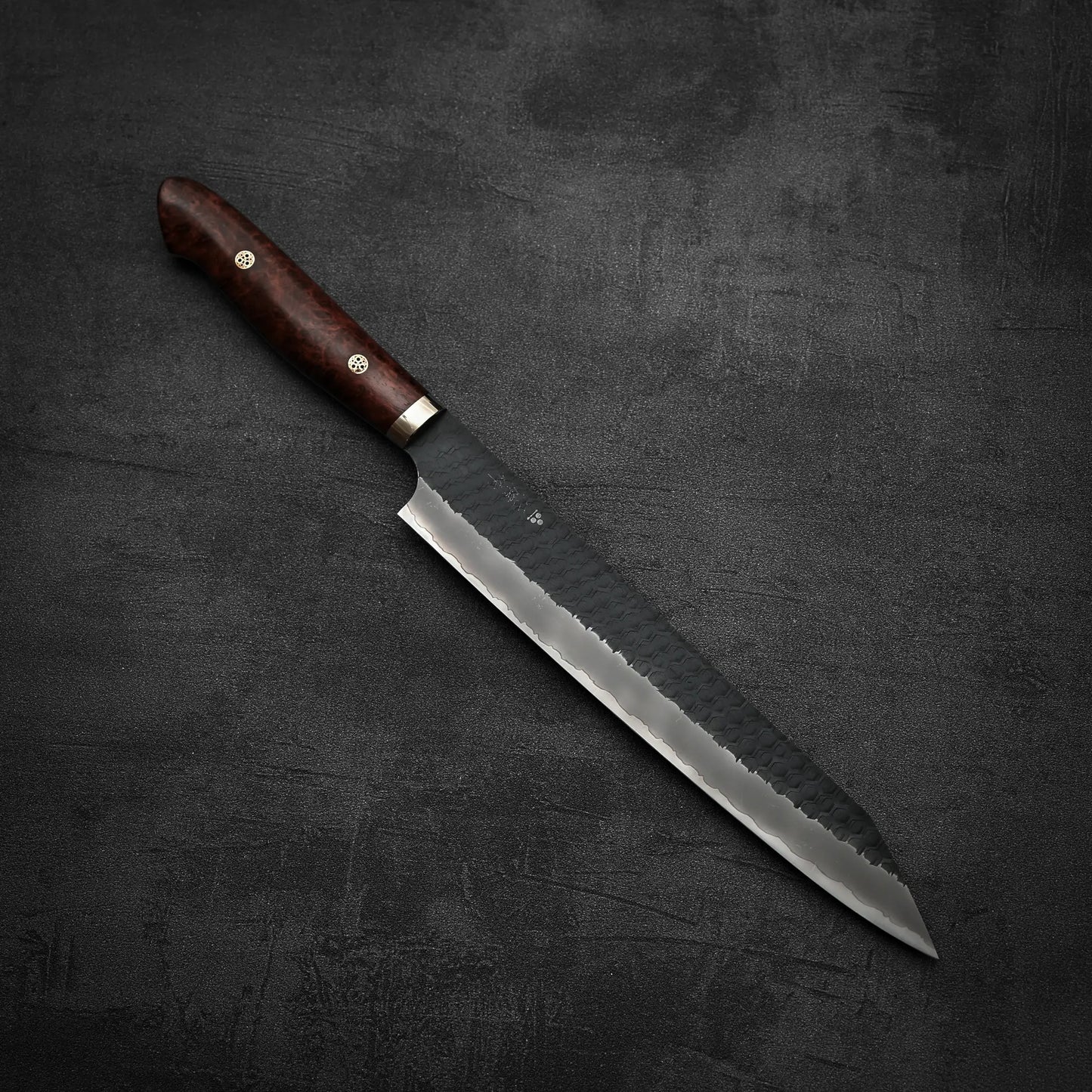 The Nigara Hamono Nigara tsuchime kurouchi SG2 kiritsuke sujihiki 255mm, featuring a dark wooden handle and hammered SG2 stainless steel blade, rests on a dark, textured surface.