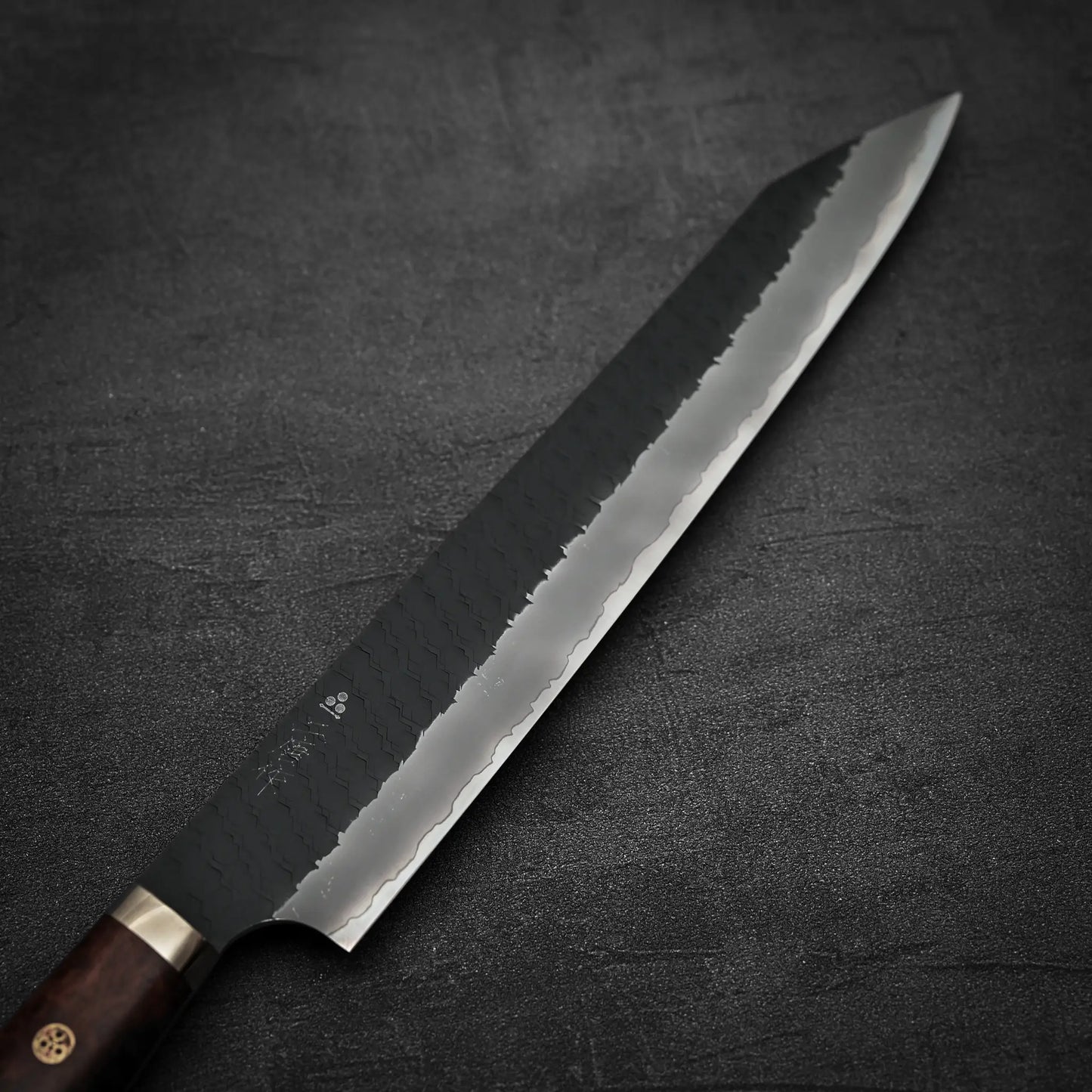 The Nigara Hamono Nigara tsuchime kurouchi SG2 kiritsuke sujihiki 255mm, featuring a hammered SG2 stainless steel blade and wooden handle, rests on a dark textured surface with its sharp, pointed tip and distinctive patterned finish on display.