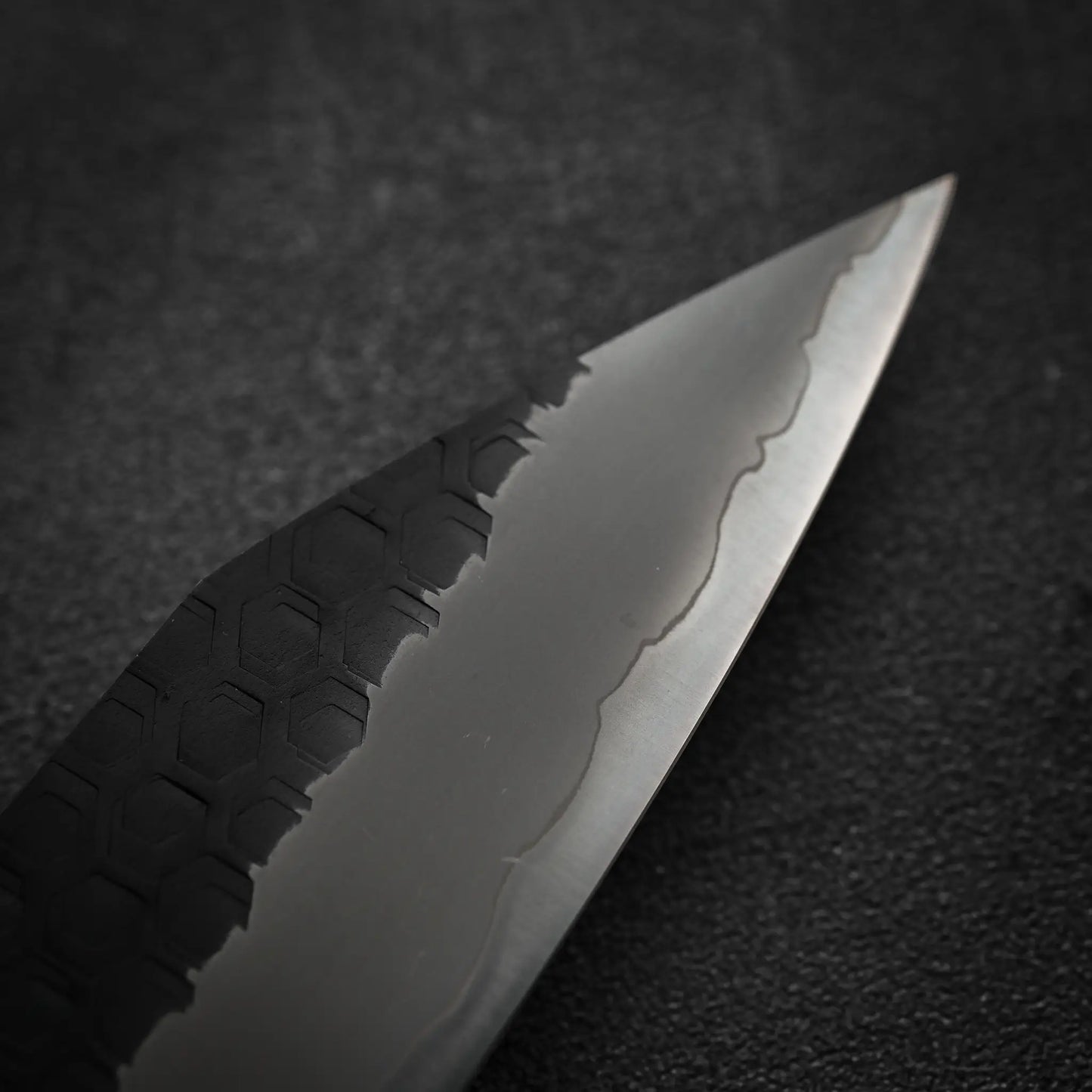 Close-up of the Nigara Hamono Nigara tsuchime kurouchi SG2 kiritsuke sujihiki 255mm blade, highlighting its sharp SG2 stainless steel edge, tsuchime kurouchi finish, and wavy line near the edge against a dark, textured background.