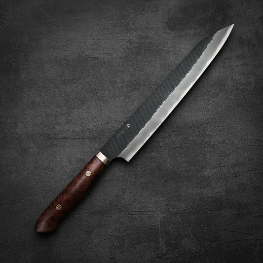 The Nigara Hamono Nigara tsuchime kurouchi SG2 kiritsuke sujihiki 255mm, featuring a hammered finish and polished wooden handle, is displayed diagonally on a textured dark gray surface.