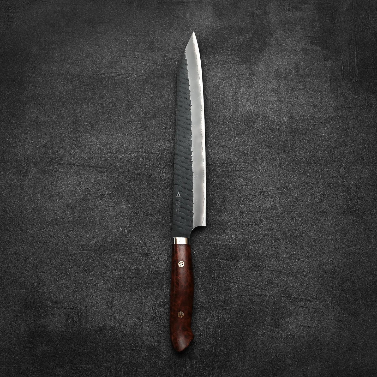 The Nigara Hamono Nigara tsuchime kurouchi SG2 kiritsuke sujihiki 255mm, featuring a polished wooden handle, rests on a dark, textured surface.