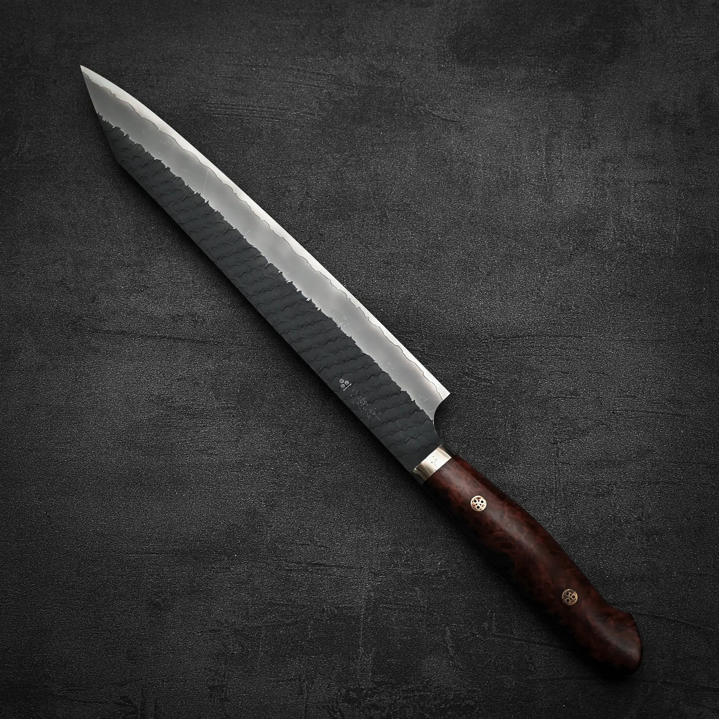 The Nigara Hamono Nigara tsuchime kurouchi SG2 kiritsuke sujihiki 255mm knife with a hammered finish and polished wooden handle lies on a textured dark gray surface.