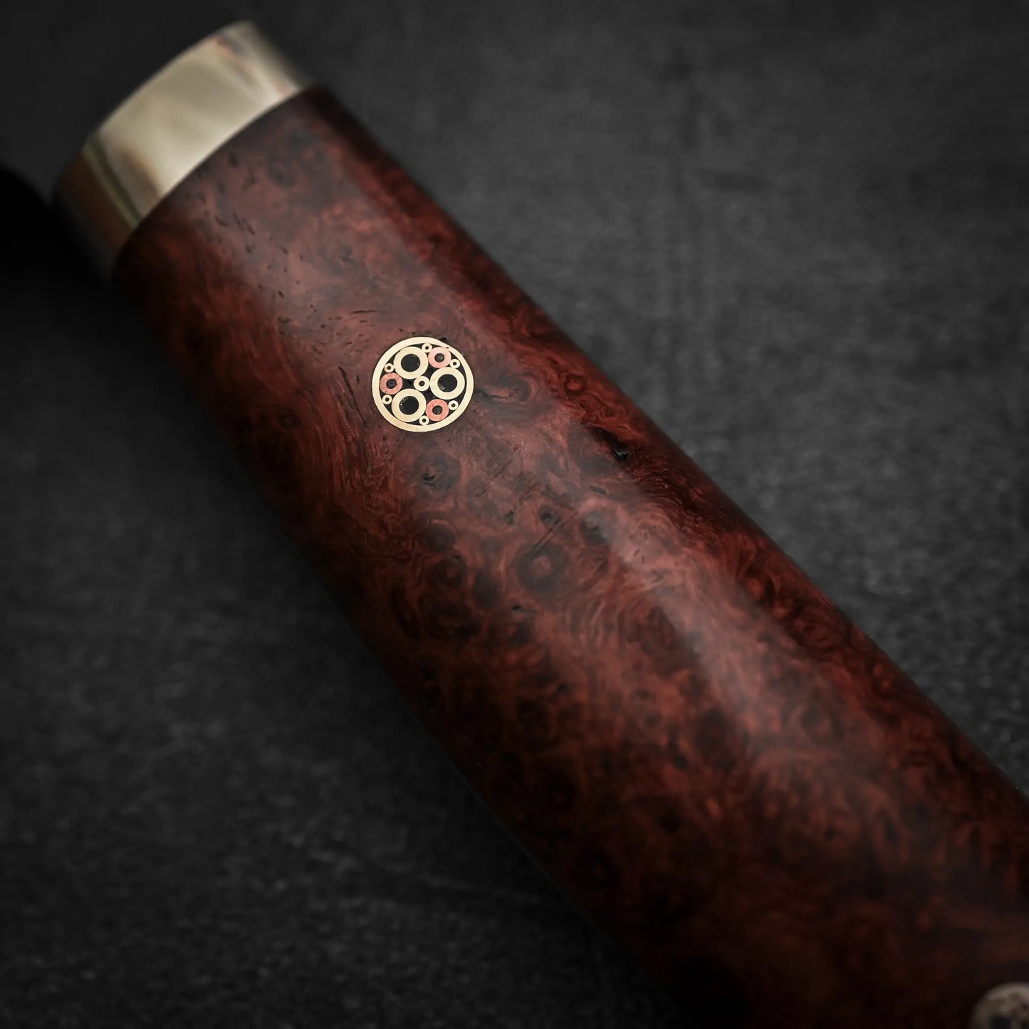 Close-up of the Nigara Hamono Nigara tsuchime kurouchi SG2 kiritsuke sujihiki 255mm knife handle, crafted from dark polished wood with a circular mosaic pin of small metal rings, set against a dark textured background.