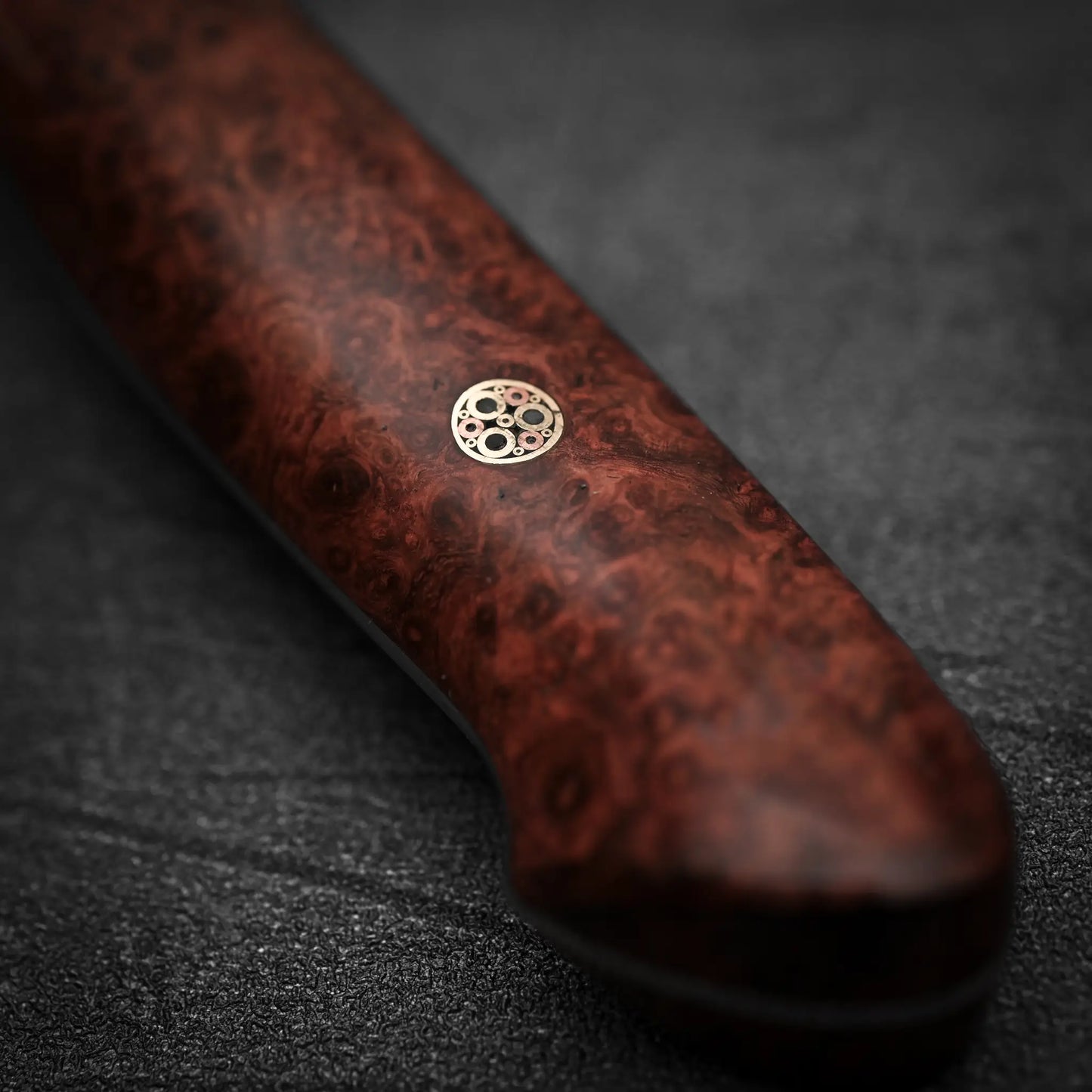 Close-up of the wooden handle with a circular mosaic pin on a Nigara Hamono Nigara tsuchime kurouchi SG2 kiritsuke sujihiki 255mm Japanese slicing knife, crafted from SG2 stainless steel, shown on a textured dark surface.