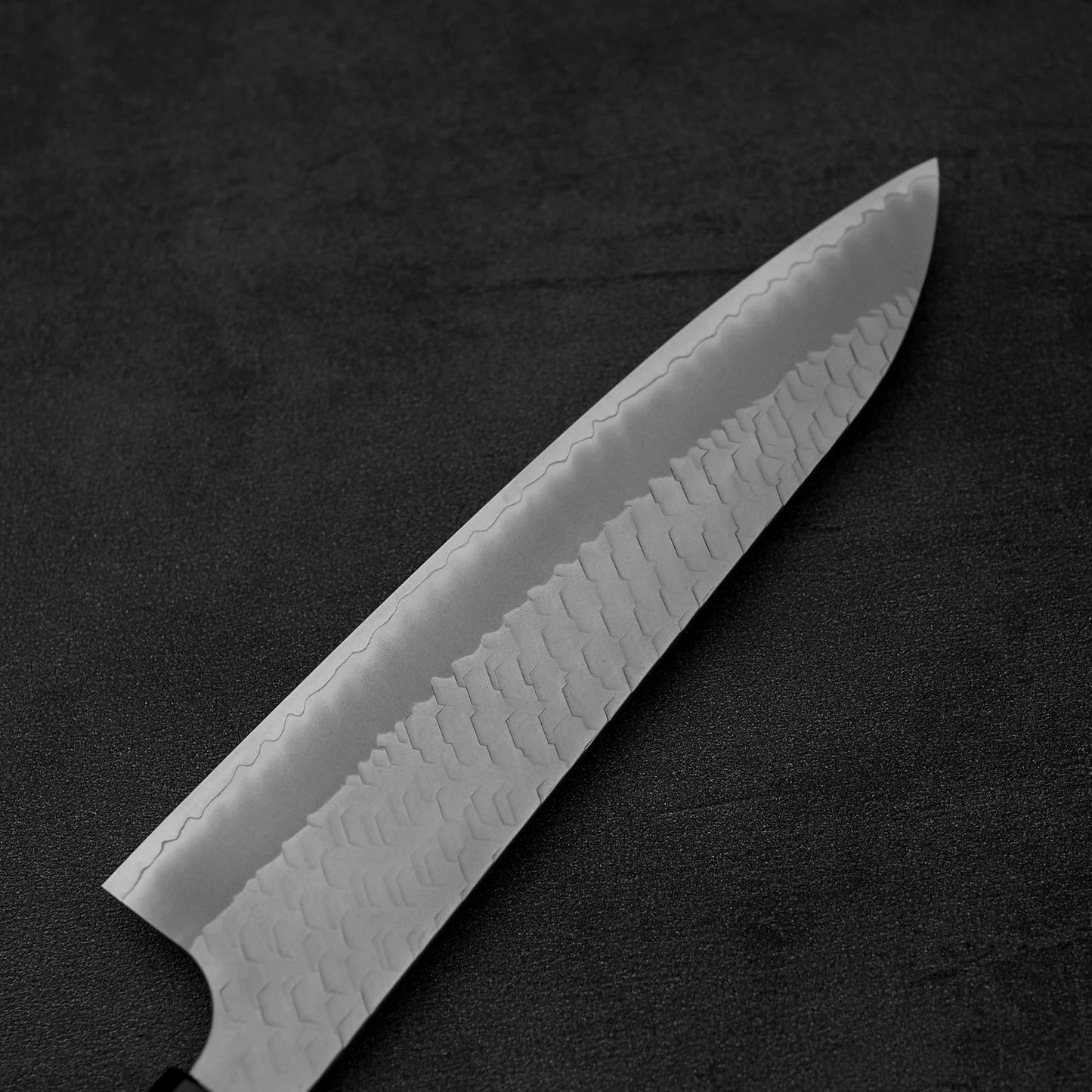 A close-up of the Nigara Hamono Nigara tsuchime SG2 gyuto 210mm knife, featuring its textured, patterned SG2 steel blade and sharp edge, resting diagonally on a dark, textured surface to highlight its craftsmanship.
