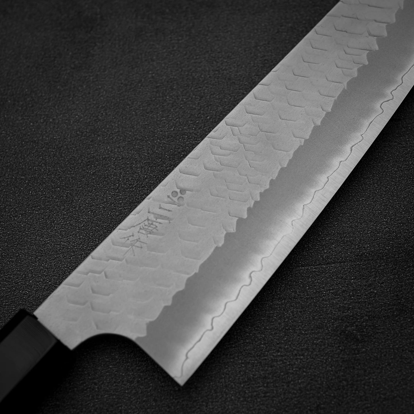 A close-up of the Nigara Hamono Nigara tsuchime SG2 gyuto 210mm, featuring a patterned SG2 steel blade with engraved characters, displayed on a dark textured surface.