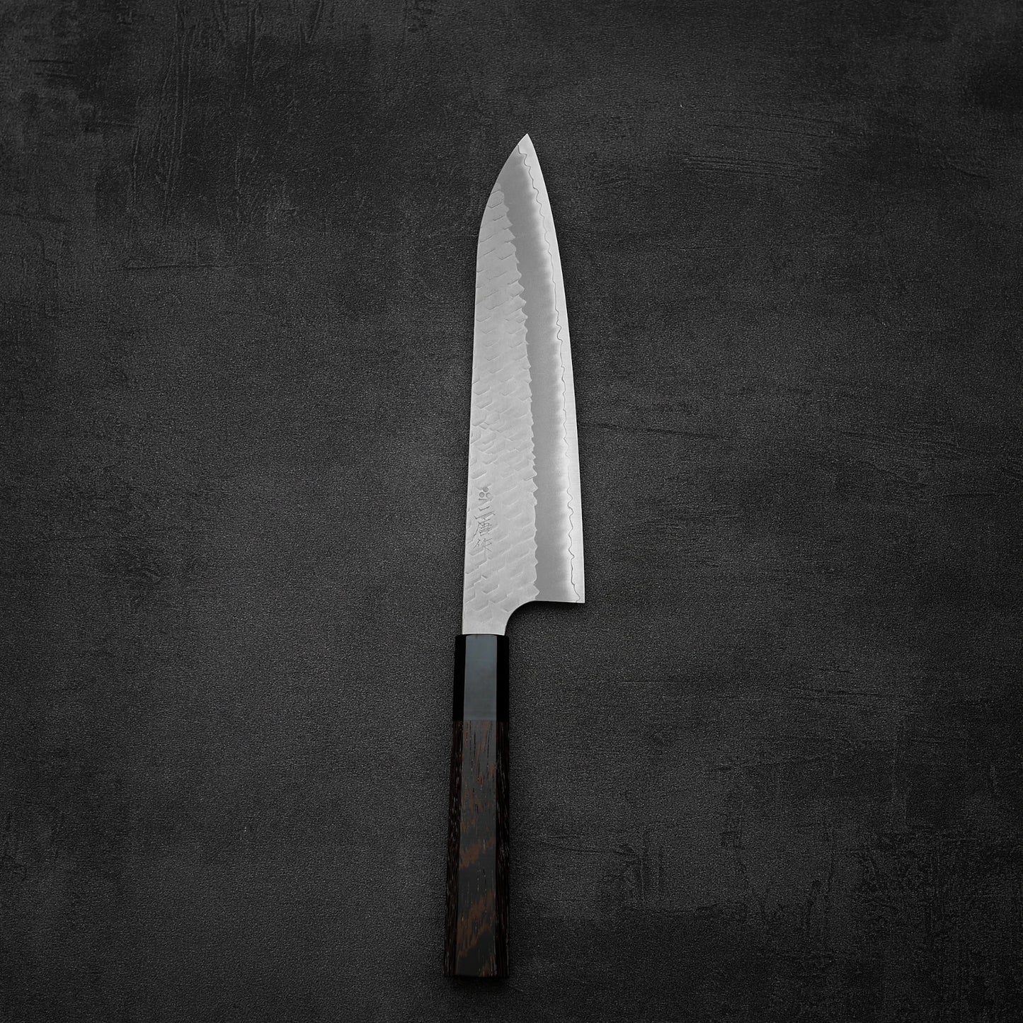 A Nigara Hamono Nigara tsuchime SG2 gyuto 210mm chefs knife with a tsuchime finish and dark wooden handle is placed on a dark textured surface.