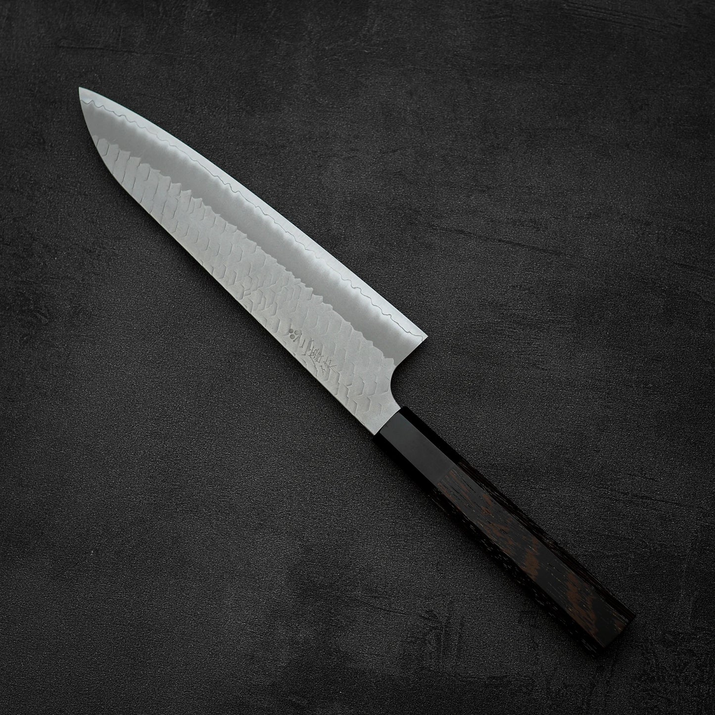 The Nigara Hamono Nigara tsuchime SG2 gyuto 210mm, with a patterned steel blade and dark wooden handle, is displayed on a black textured surface.