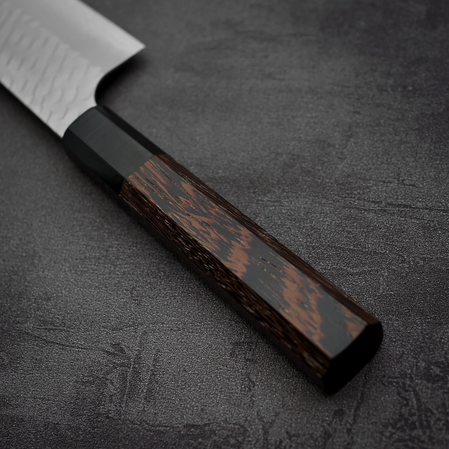A close-up of the Nigara Hamono Nigara tsuchime SG2 gyuto 210mm, showcasing its wooden handle and stainless steel blade with a tsuchime finish, resting on a dark textured surface.