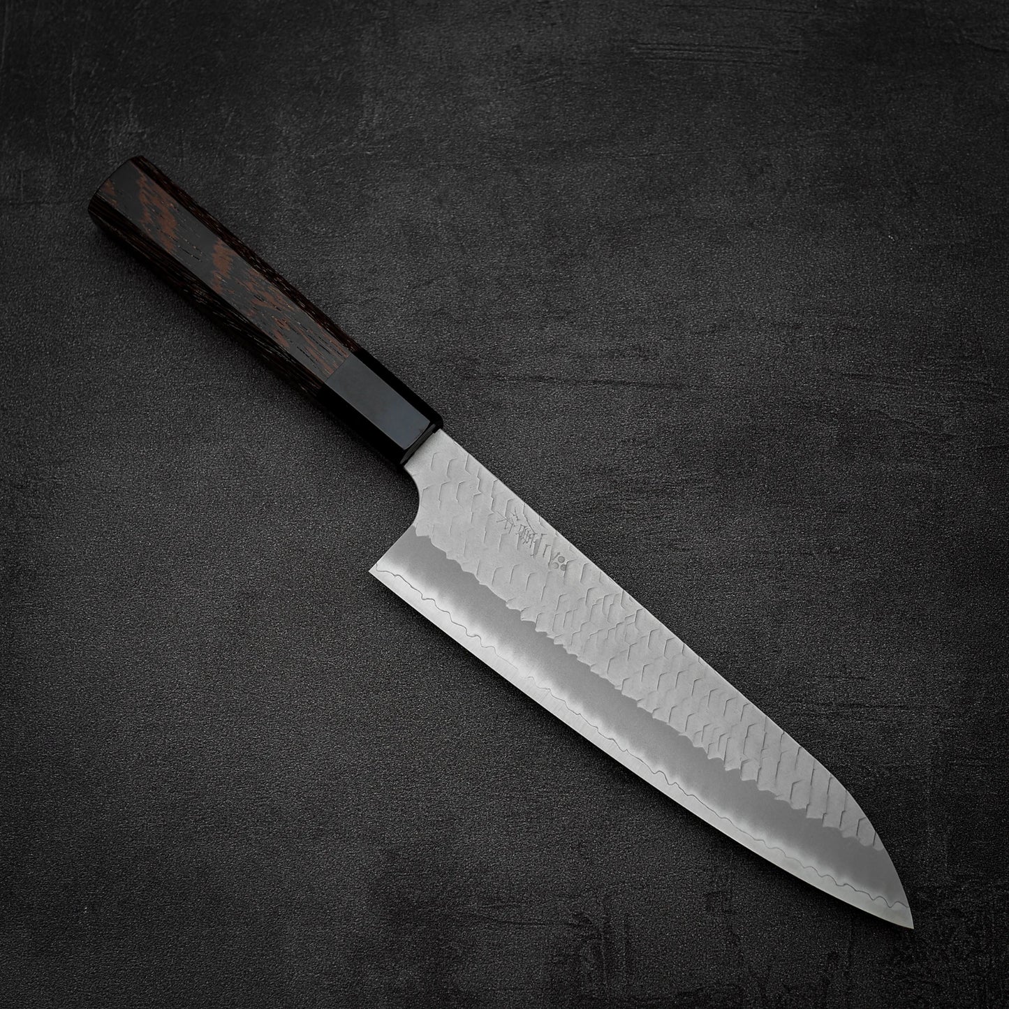 The Nigara tsuchime SG2 gyuto 210mm by Nigara Hamono, featuring a hammered steel blade and dark wooden handle, rests on a textured black surface.
