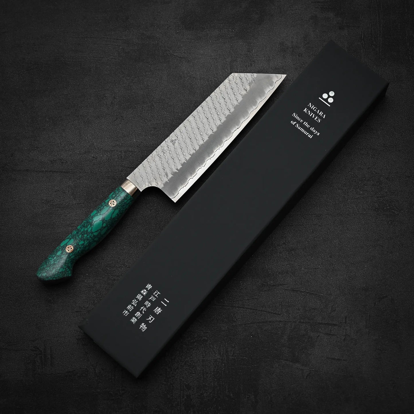 The Nigara Hamono Nigara tsuchime SG2 kiritsuke nakiri 180mm, featuring a hammered SG2 blade and green marbled handle, is shown beside a black box with white Japanese and English text on a dark, textured backdrop.