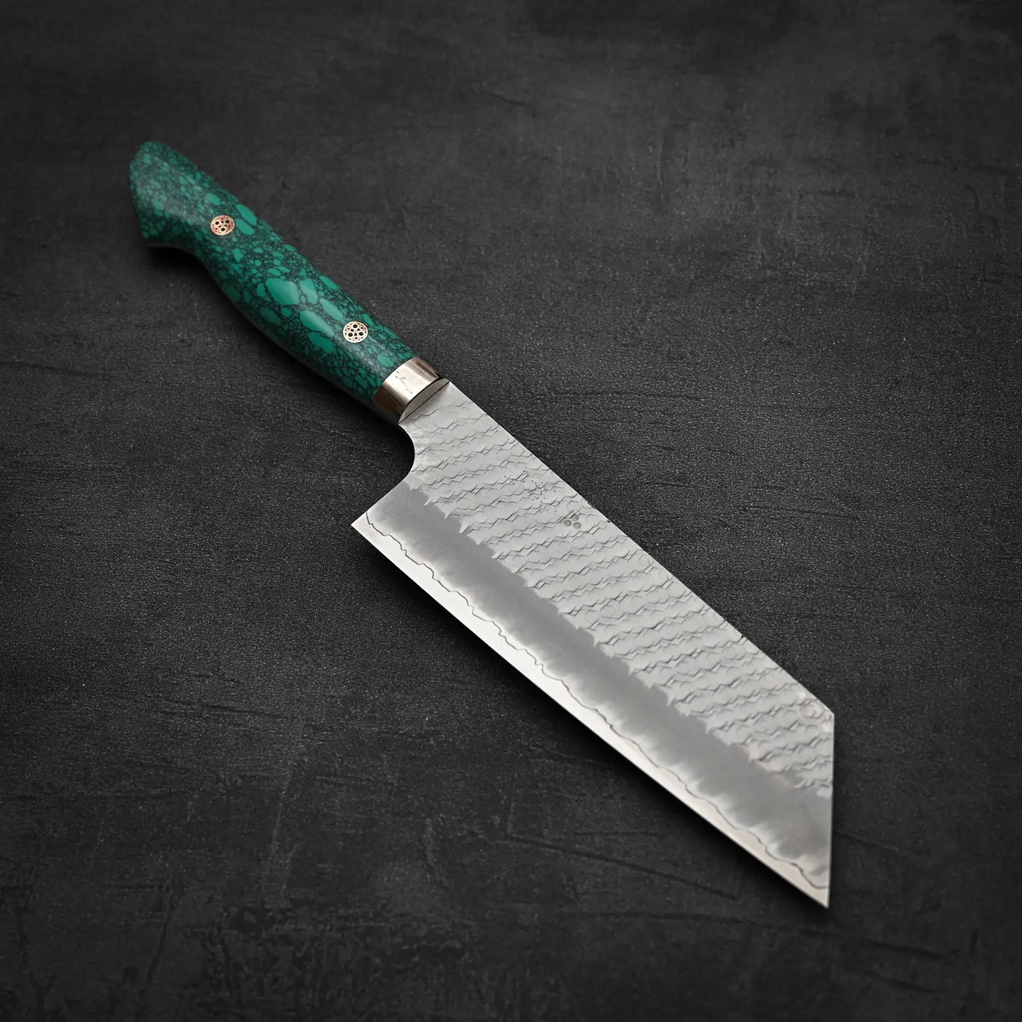 The Nigara Hamono Nigara tsuchime SG2 kiritsuke nakiri 180mm, featuring an SG2 patterned blade and green marbled handle, rests on a dark textured surface—ideal for precise vegetable prep with its straight edge and angled tip.