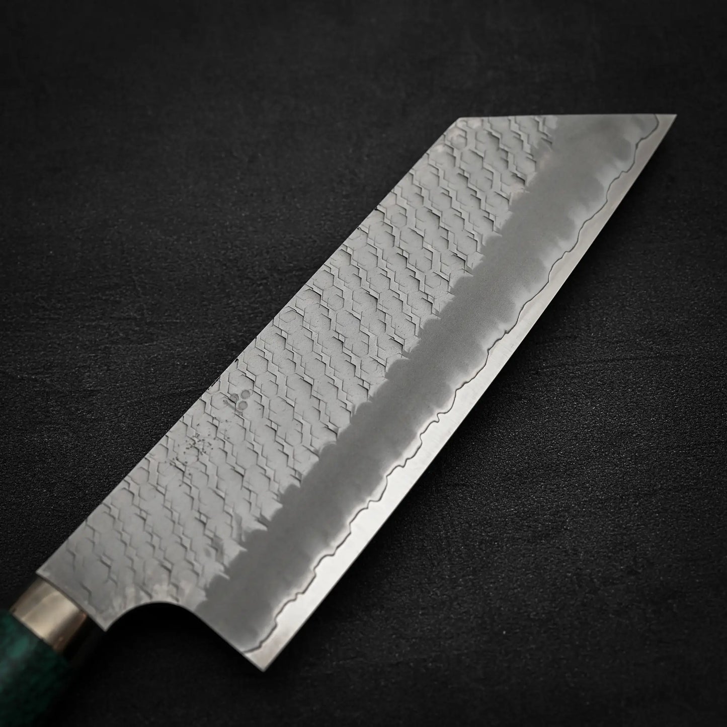 A close-up of the Nigara Hamono Nigara tsuchime SG2 kiritsuke nakiri 180mm, showcasing its patterned SG2 stainless steel blade and green handle, set against a dark, textured background.