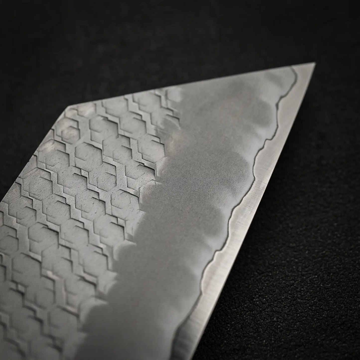 Close-up of the Nigara Hamono Nigara tsuchime SG2 kiritsuke nakiri 180mm blade, featuring intricate geometric patterns near the spine and a wavy edge line against a dark, textured background.